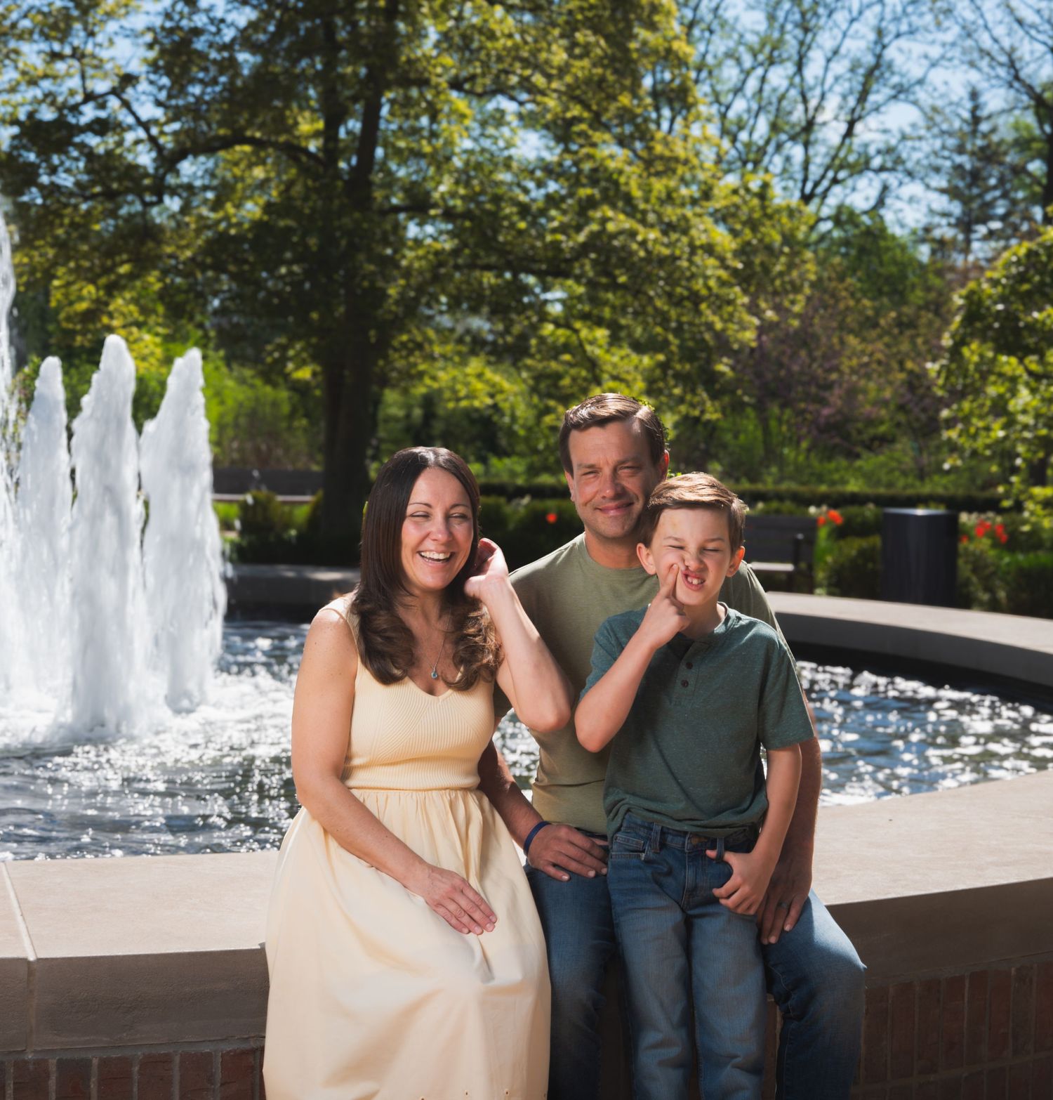 A smiling family of three poses by a brick fountain surrounded by lush green trees on a sunny day.