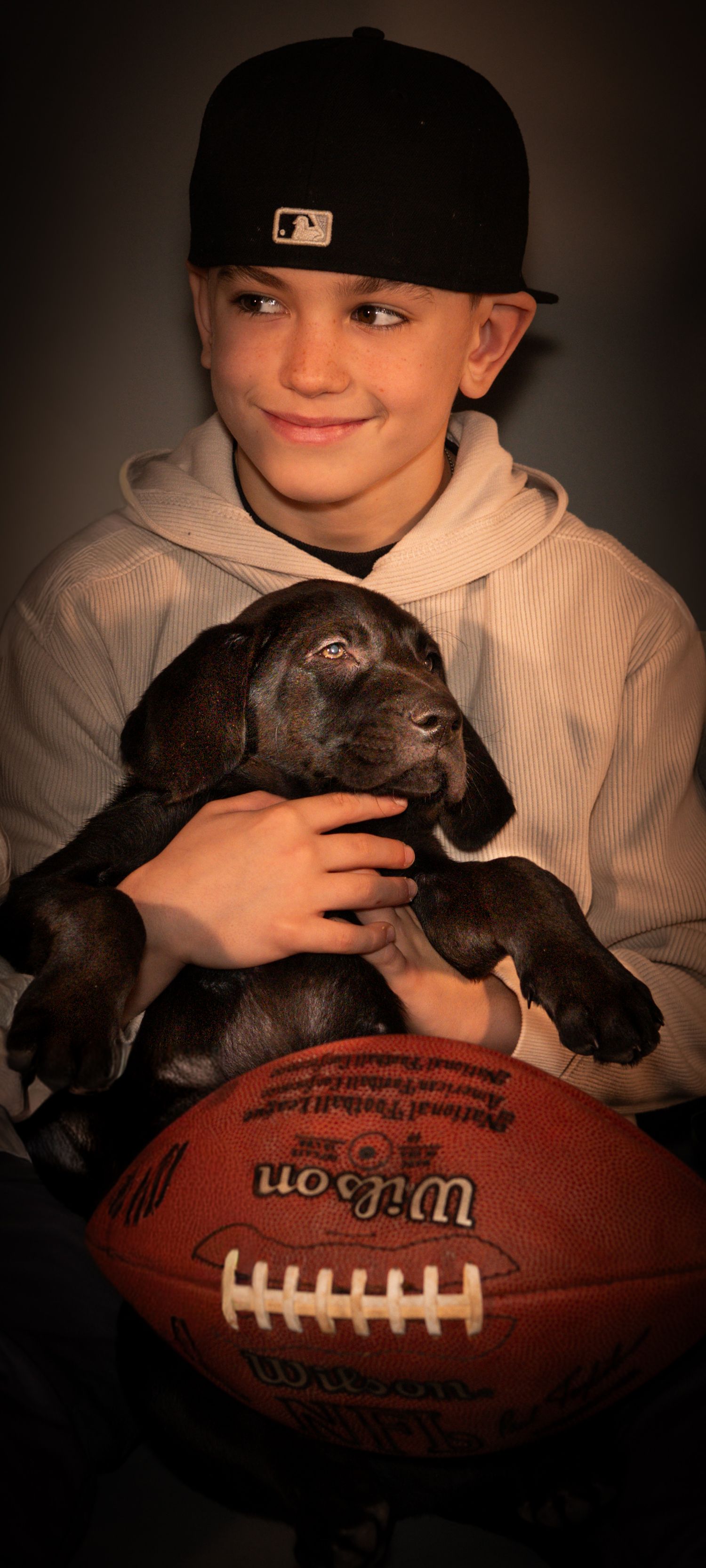 A small black puppy rests on a football while someone in a baseball cap and hoodie holds them in a dimly lit setting.