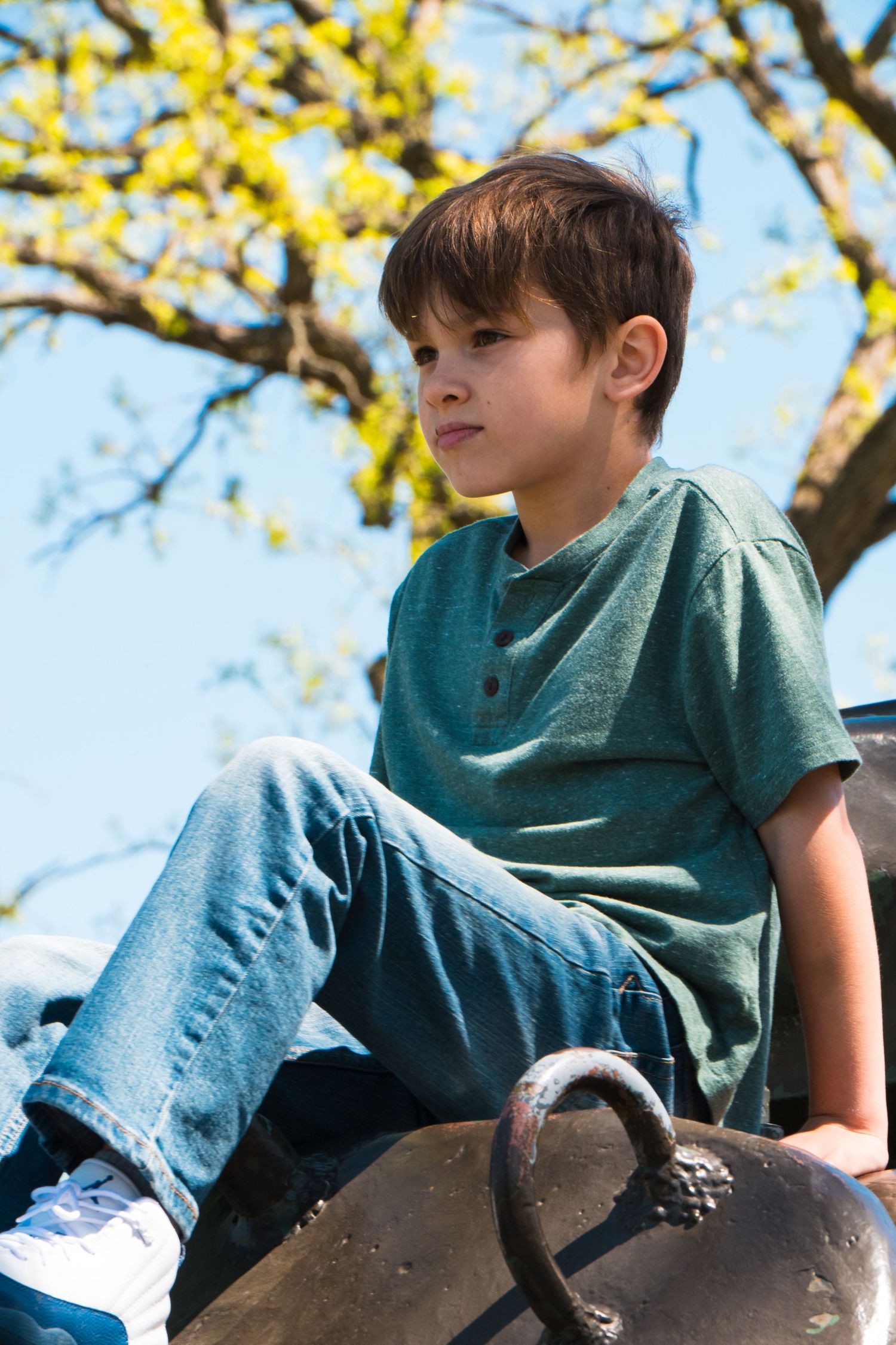 A young boy in a teal shirt and jeans sits outdoors, gazing thoughtfully with a tree and blue sky behind him.