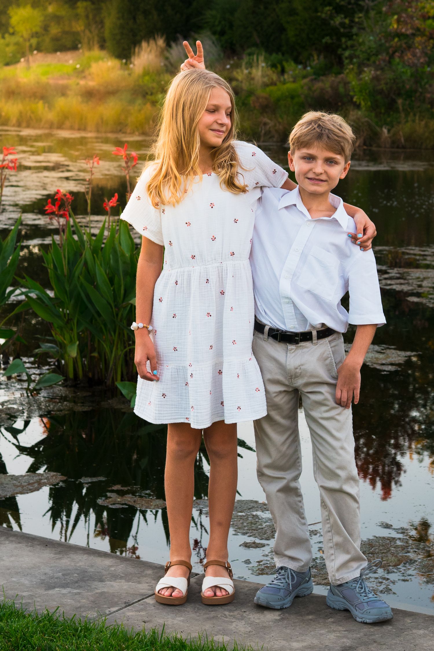 Two smiling children, a girl in a white floral dress and a boy in a white shirt and khaki pants, pose beside a pond with red flowers.