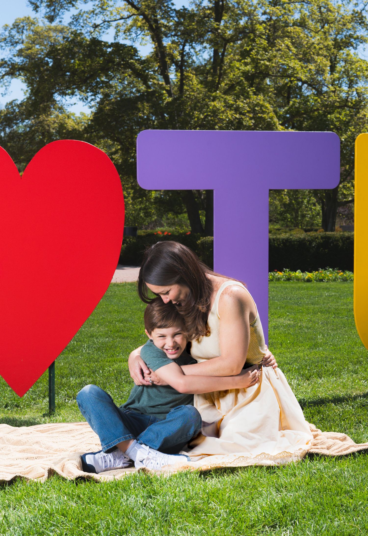 A mother hugs her smiling child on a blanket in a park beside colorful large letter decorations on green grass.