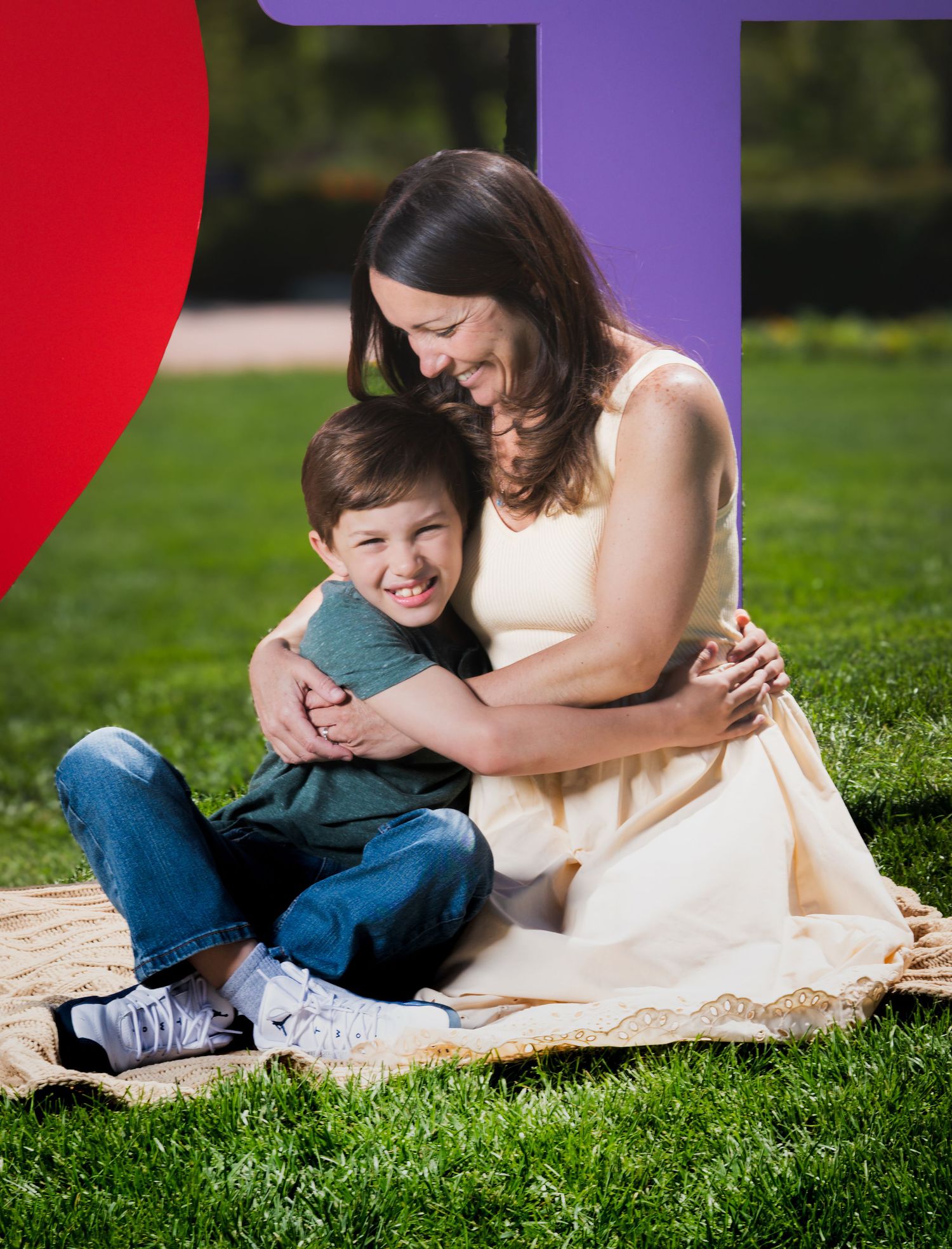 A woman and young boy share a warm embrace while sitting on a blanket in a lush green park setting.