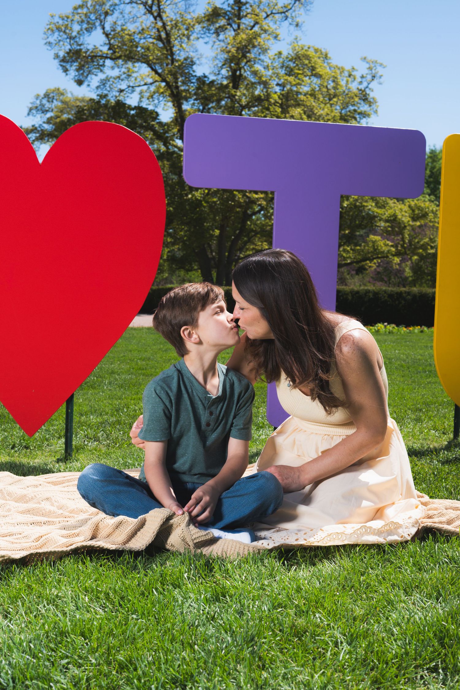 A boy kisses his pregnant mother on a blanket in a park surrounded by colorful letter and heart decorations.