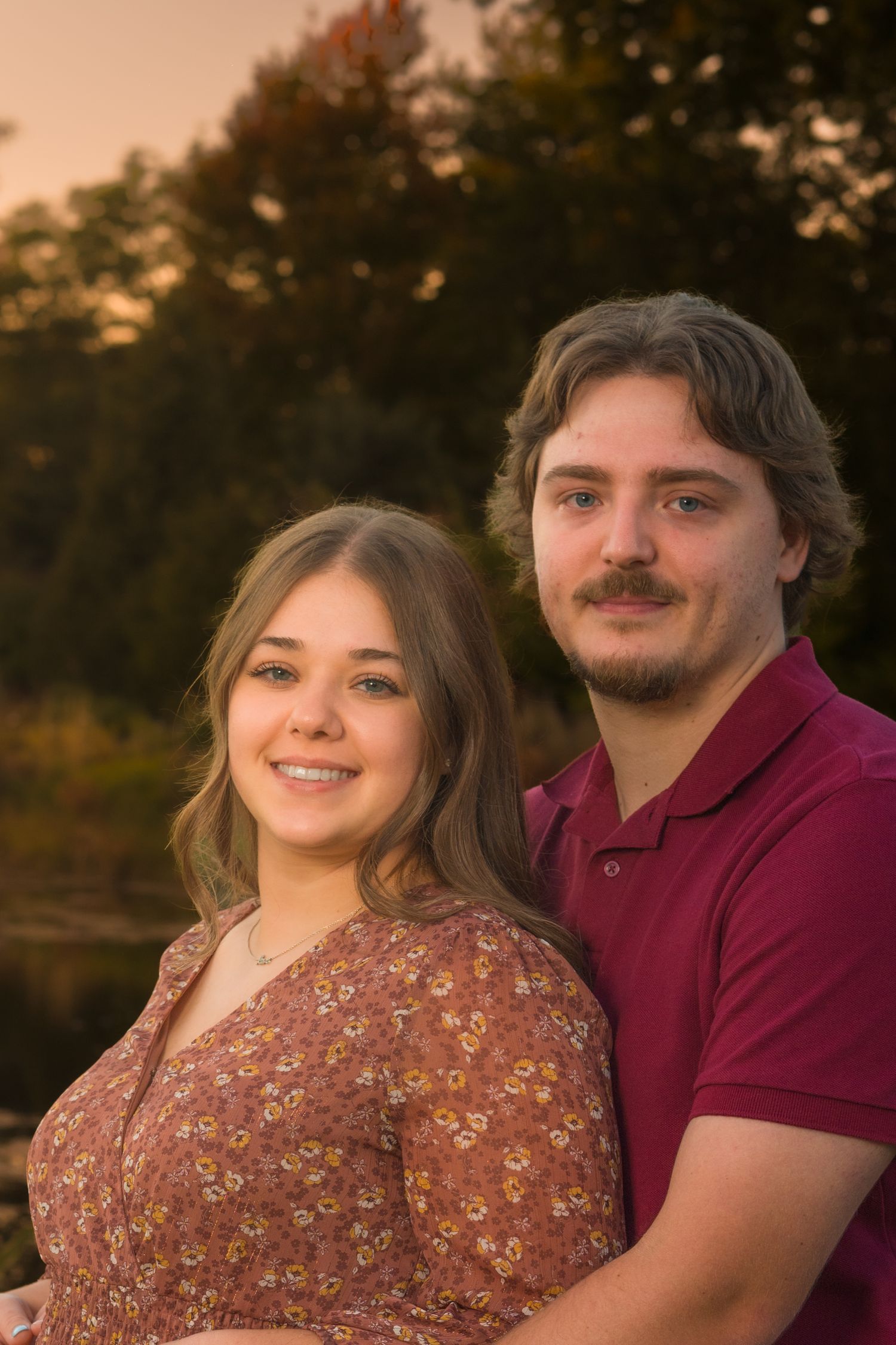 A smiling young woman in a floral top and a bearded man in a maroon polo pose close together outdoors near a lake at golden sunset.