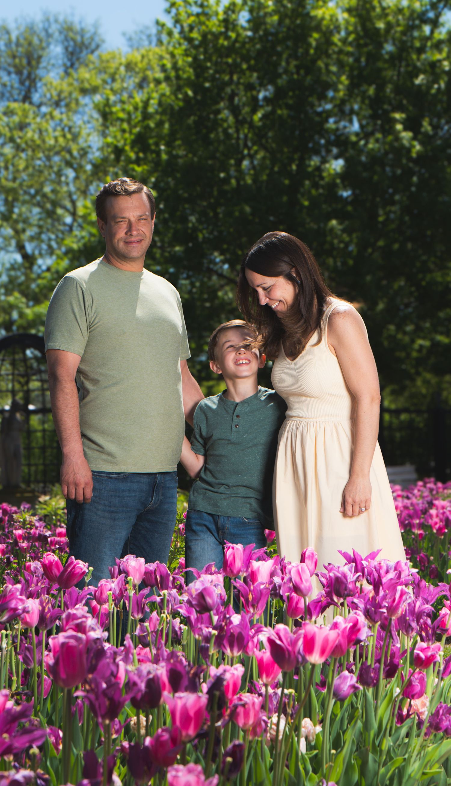 Family of three smiling among vibrant pink tulips in a sunny garden, with lush green trees in the background.