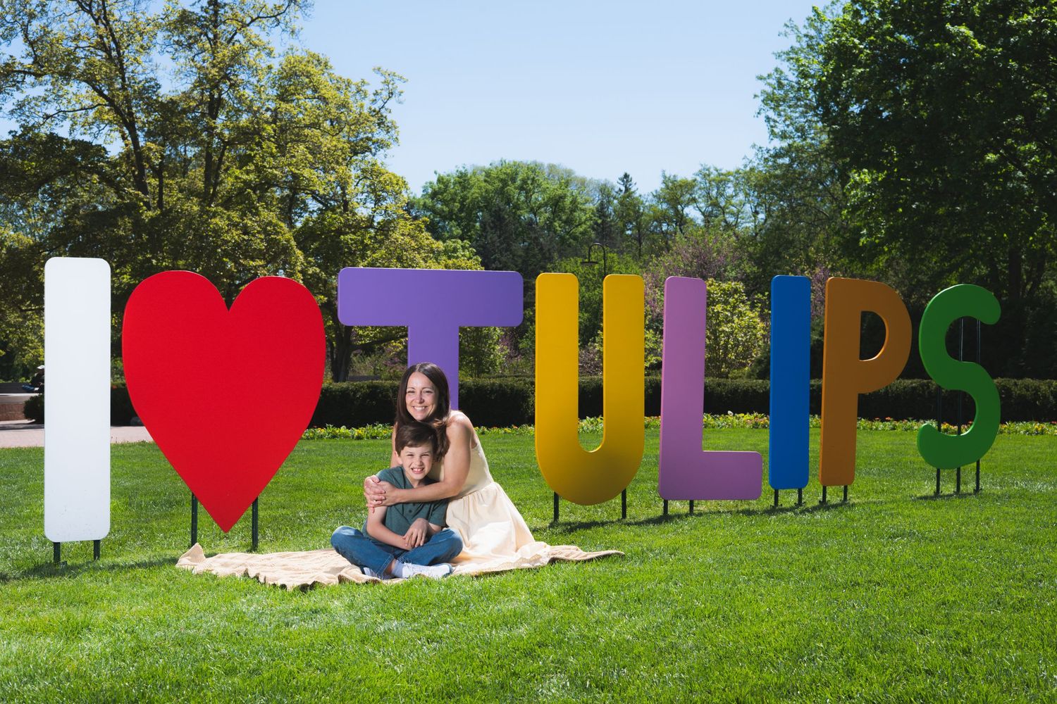 Woman and child sitting on a blanket in front of colorful I Love Tulips sign in a sunny green park.