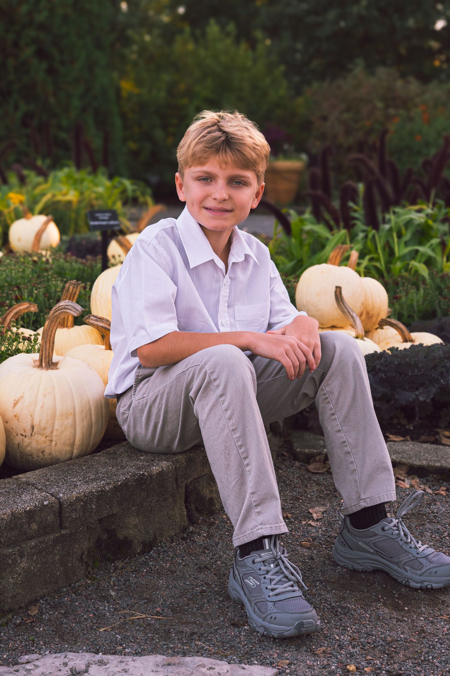 A smiling boy in a white button-up shirt and khaki pants sits on a stone ledge beside white pumpkins in a autumn garden.