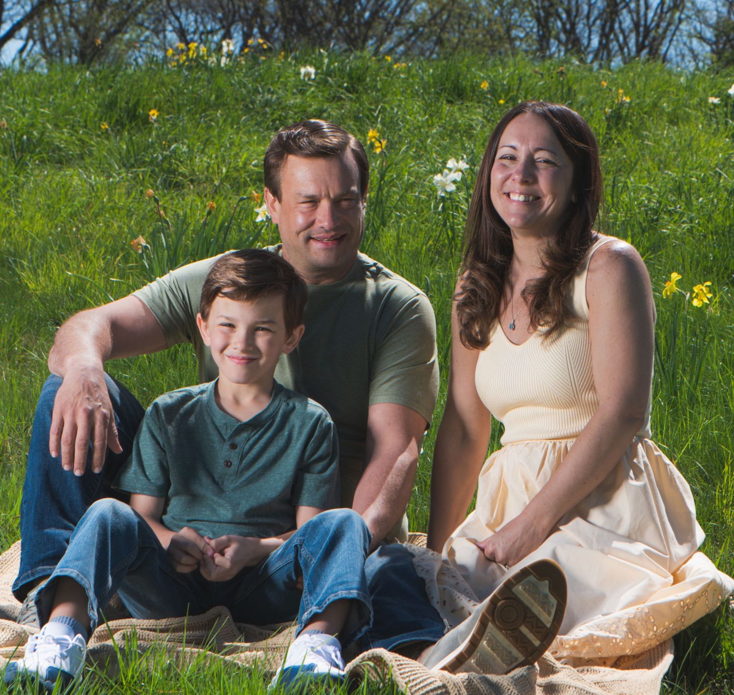 Family of three smiling and sitting on a blanket in a green meadow with wildflowers on a sunny day.