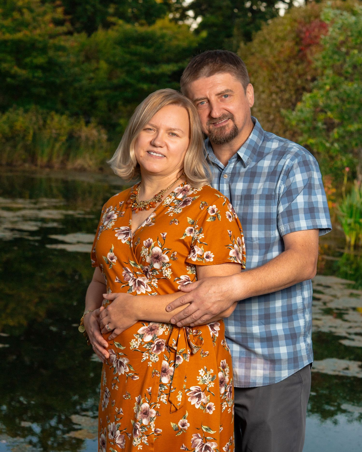 A smiling couple embraces outdoors beside a calm pond, the woman in an orange floral dress and the man in a blue plaid shirt.