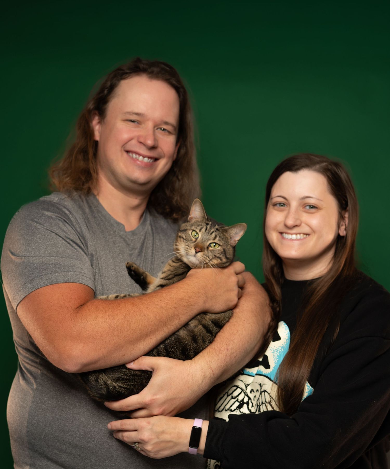 A smiling couple holds a tabby cat against a green background in a professional portrait photo.
