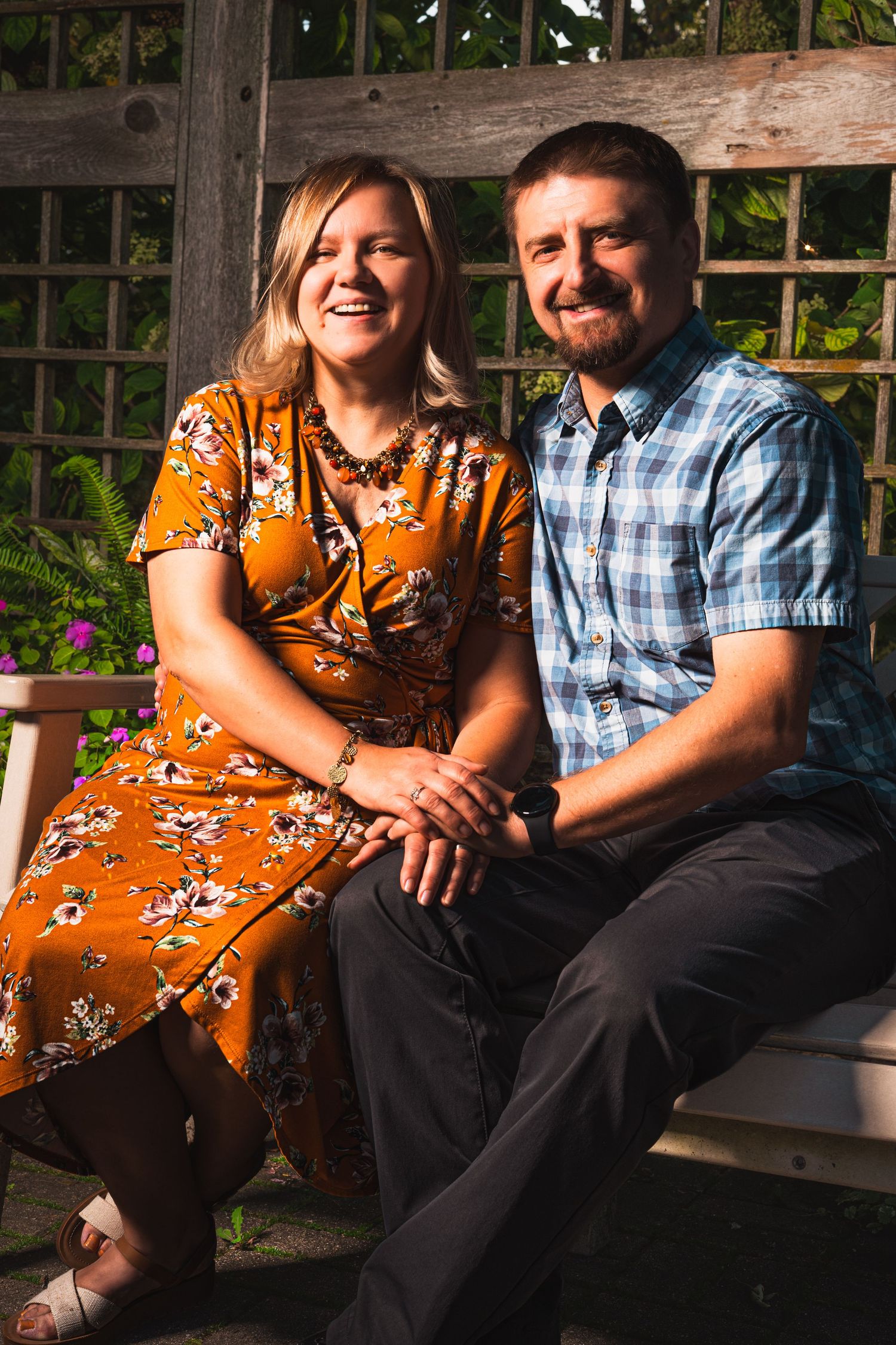 Smiling couple sitting close together on a wooden garden bench, the woman in an orange floral dress and the man in a blue plaid shirt.