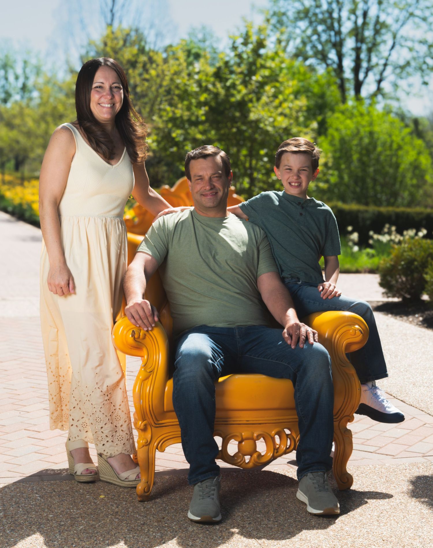 Family of three posing outdoors on sunny day, woman in white dress, man seated in yellow chair, boy standing behind.