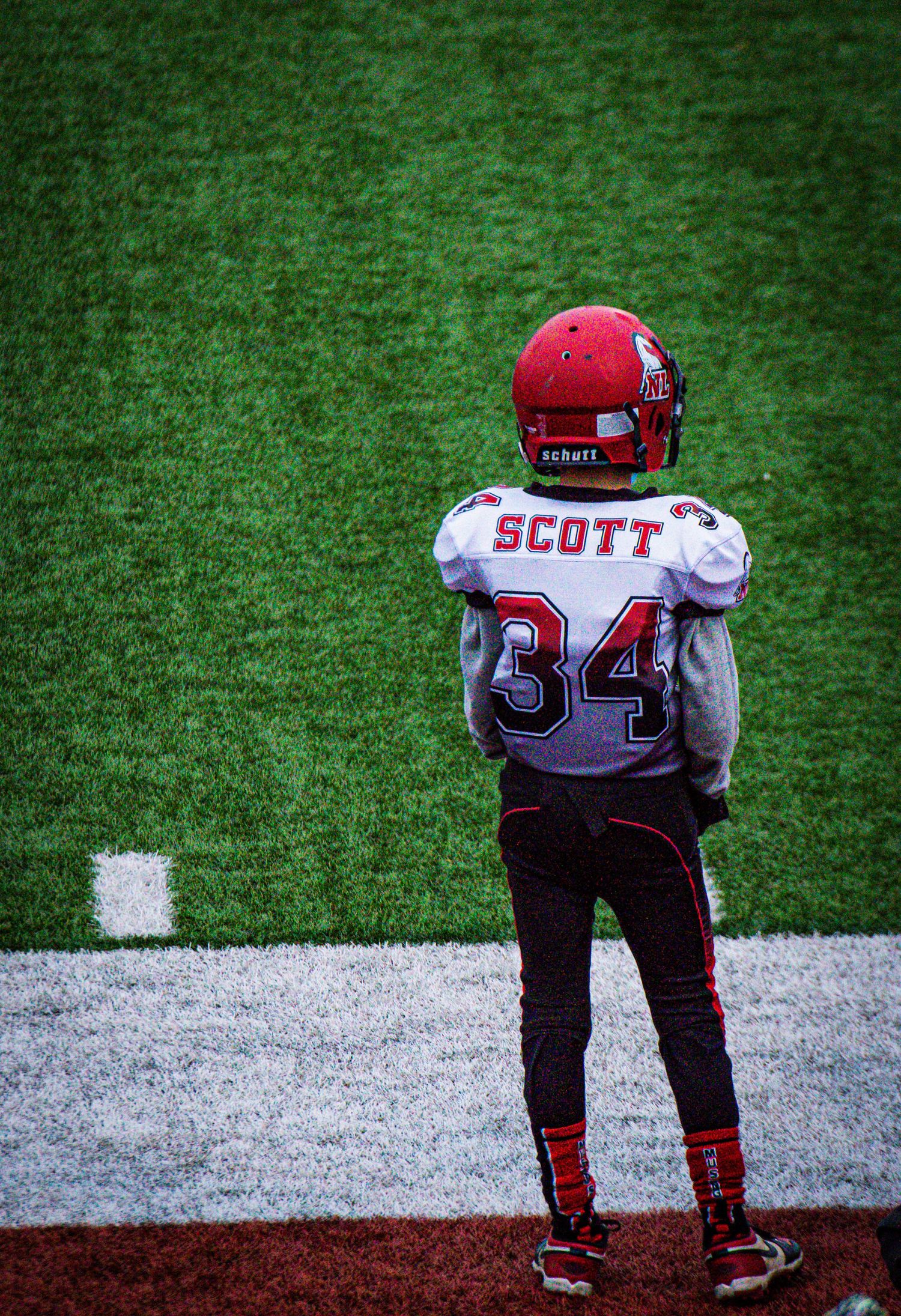 Youth baseball player in red and white uniform with 'SCOTT' on jersey stands on baseball field during game.