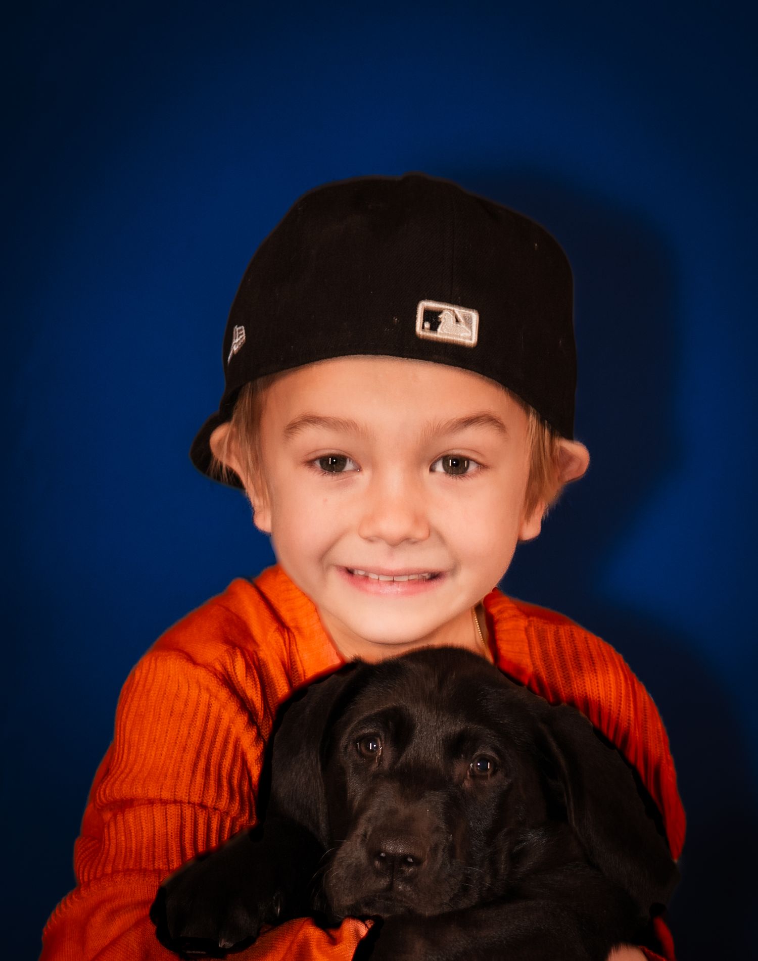 A child in an orange sweater poses with a black Labrador dog against a dark blue background.