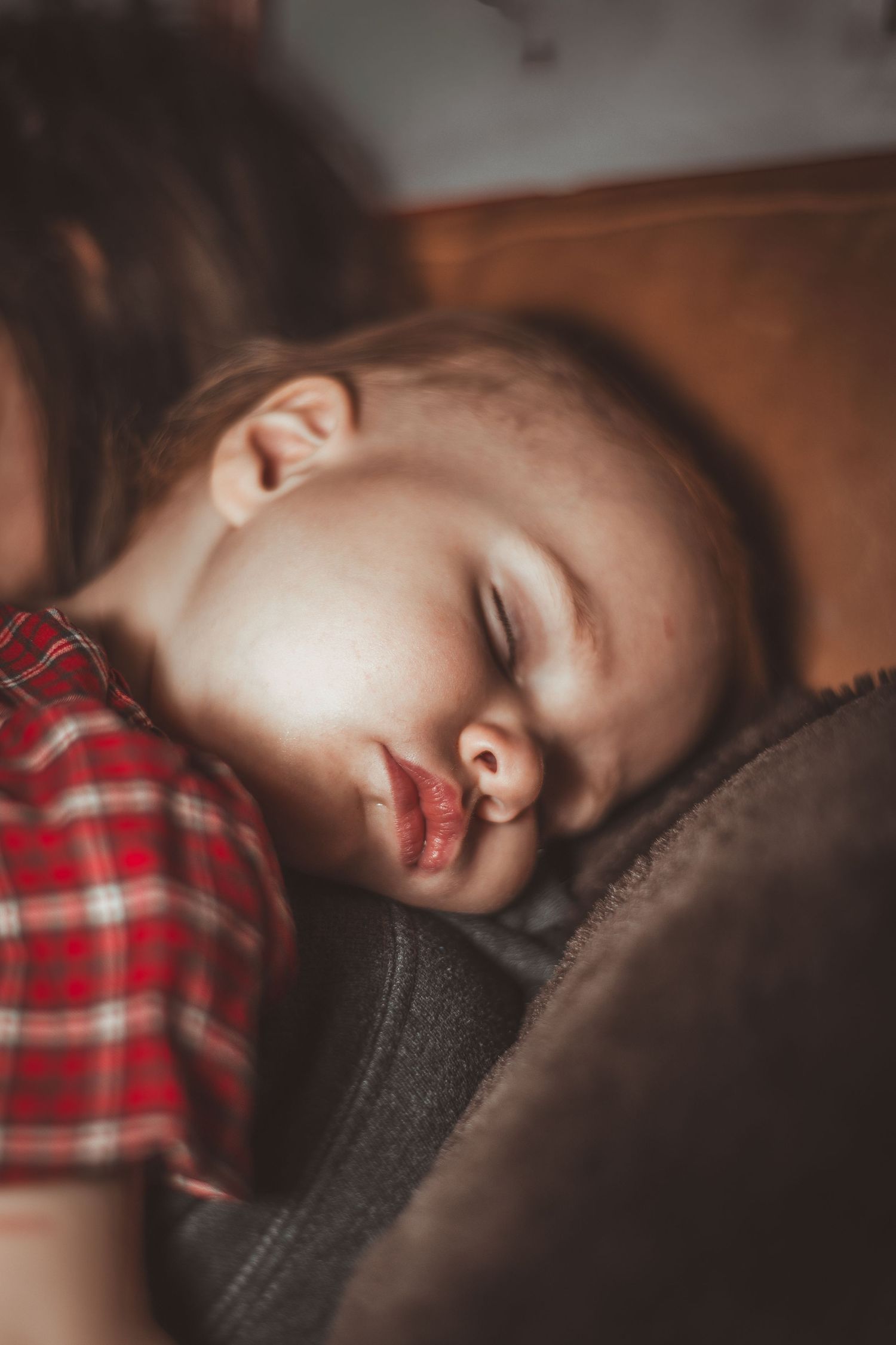A child wearing a red plaid shirt sleeps peacefully on a dark fabric surface in moody lighting.