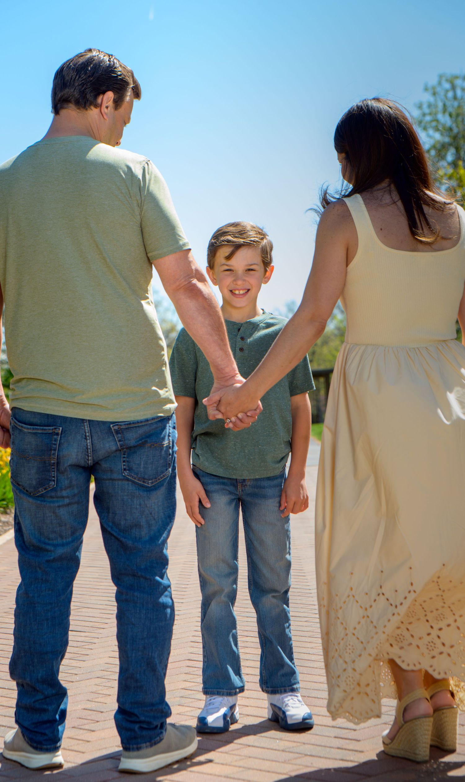 A smiling boy holds hands with his parents on a sunny brick pathway, looking back at the camera.