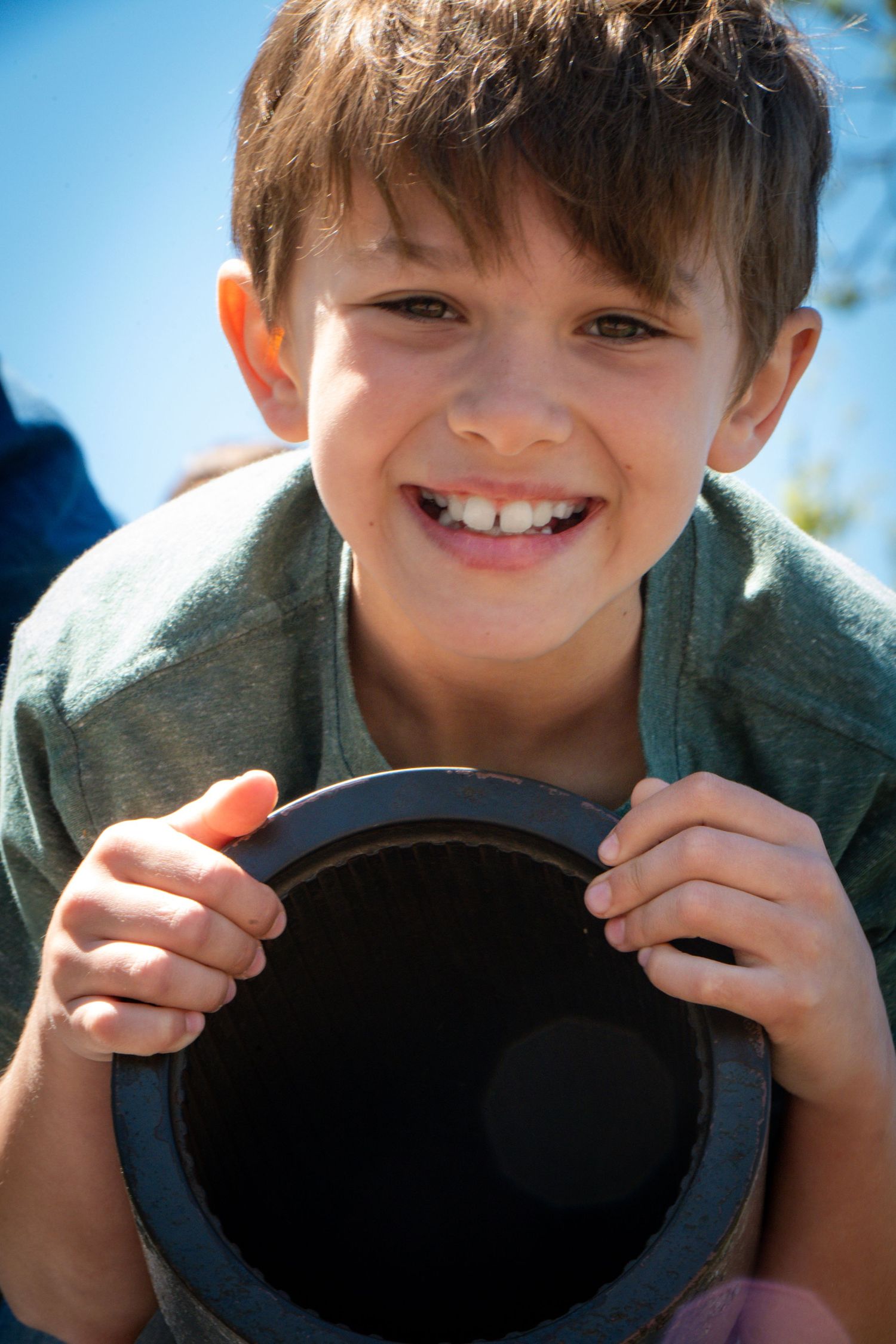 Smiling young boy holding a dark circular object outdoors on a sunny day, wearing a green shirt.