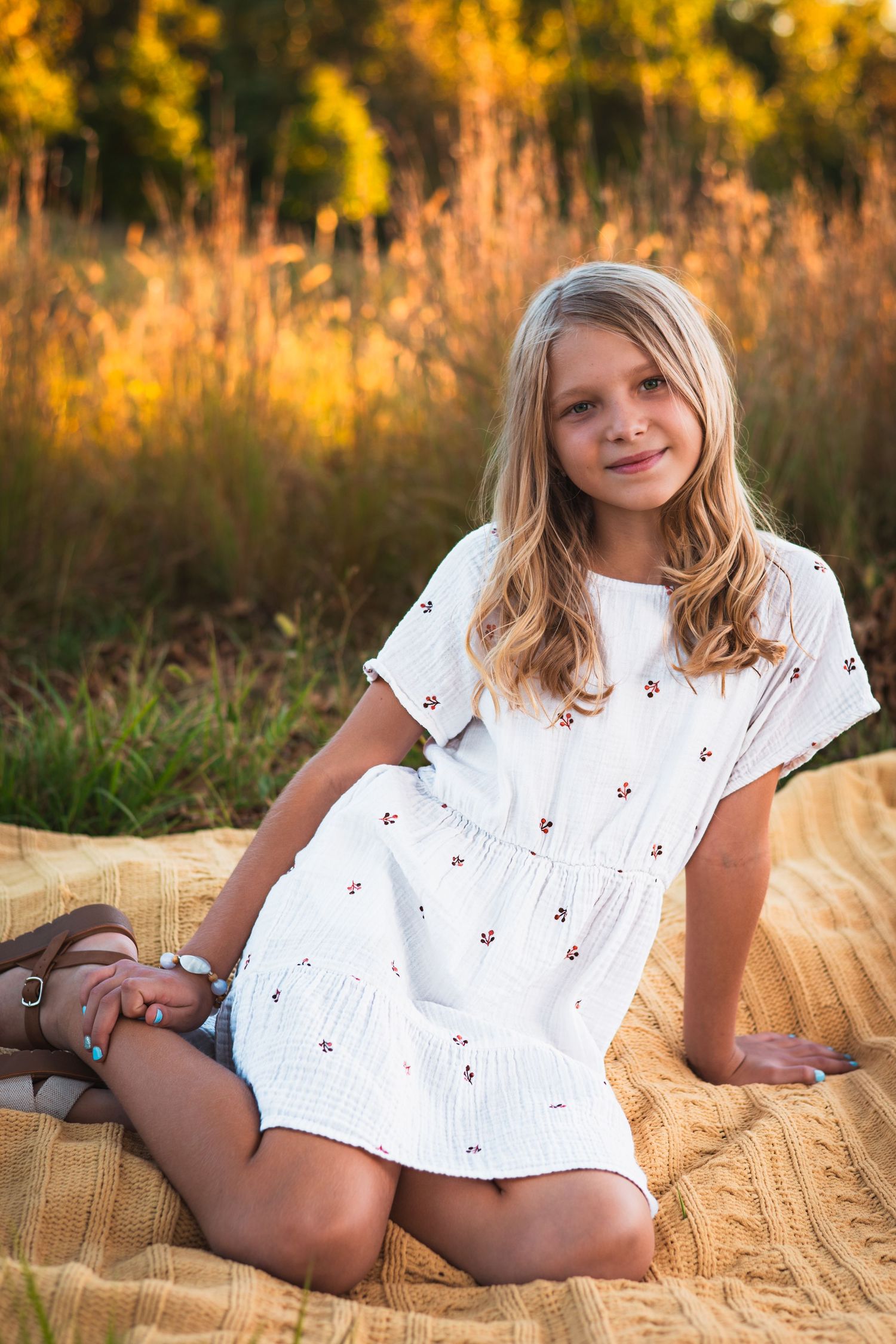 Smiling girl with long blonde hair in a white floral dress sits on a tan knit blanket in a golden sunlit field.
