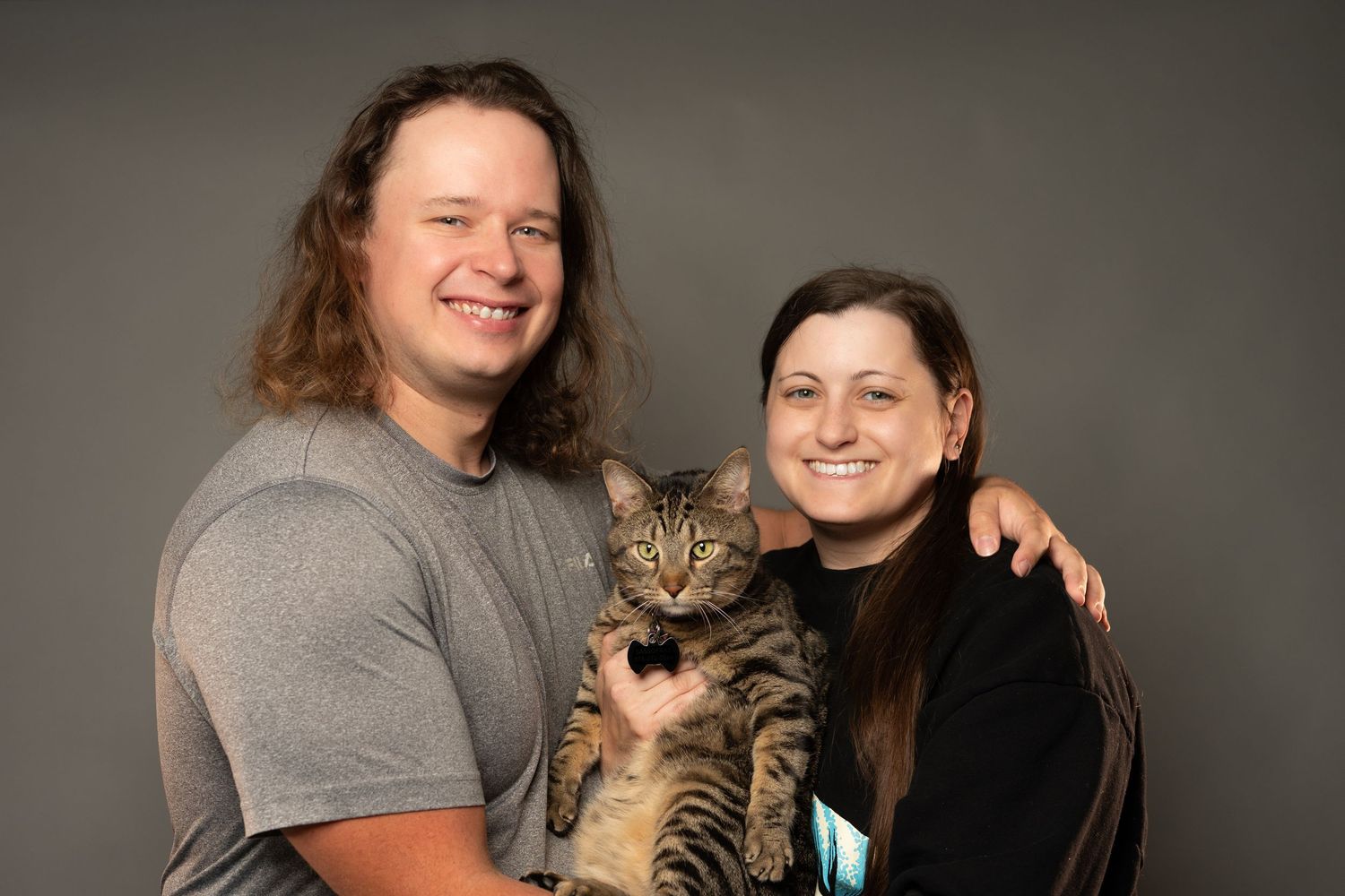 A smiling couple poses together holding a tabby cat against a gray studio background.