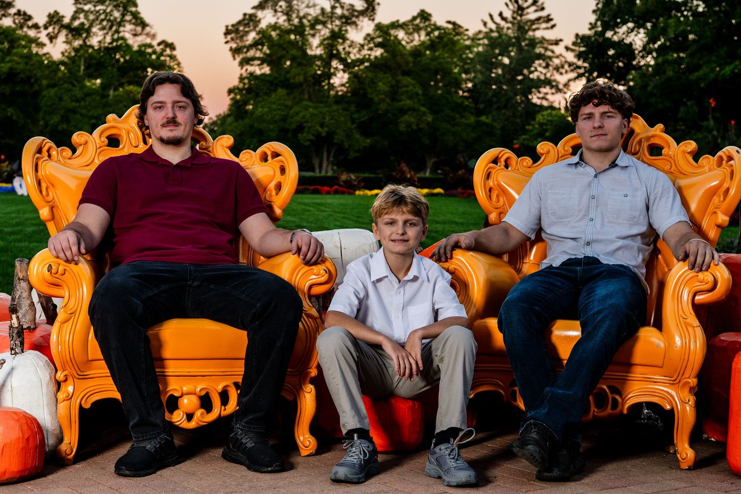 Two young men and a boy pose on ornate orange throne chairs outdoors at sunset, with green trees and a lawn behind them.