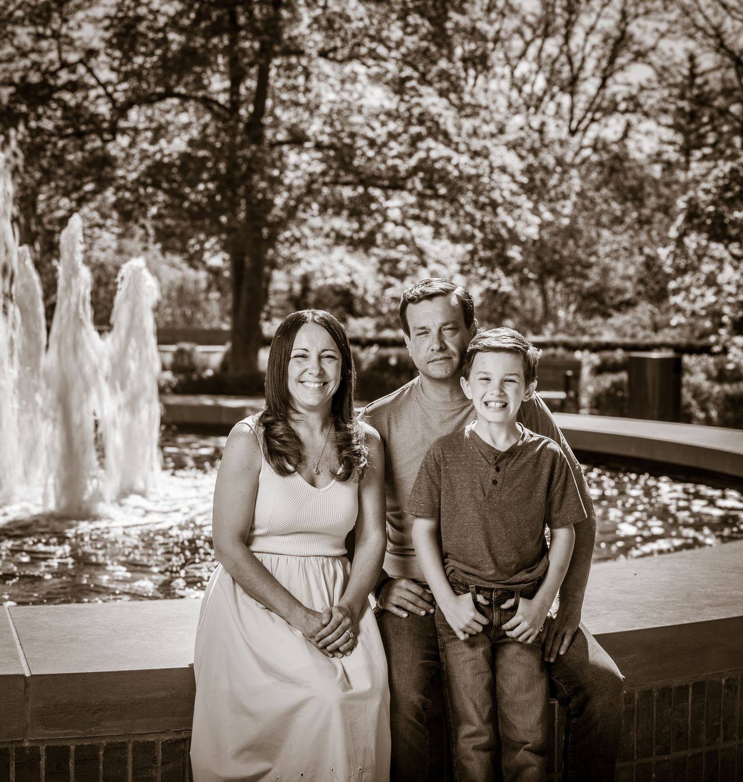 A family of three smiles together near a fountain in a lush park, captured in a vintage sepia-toned portrait.