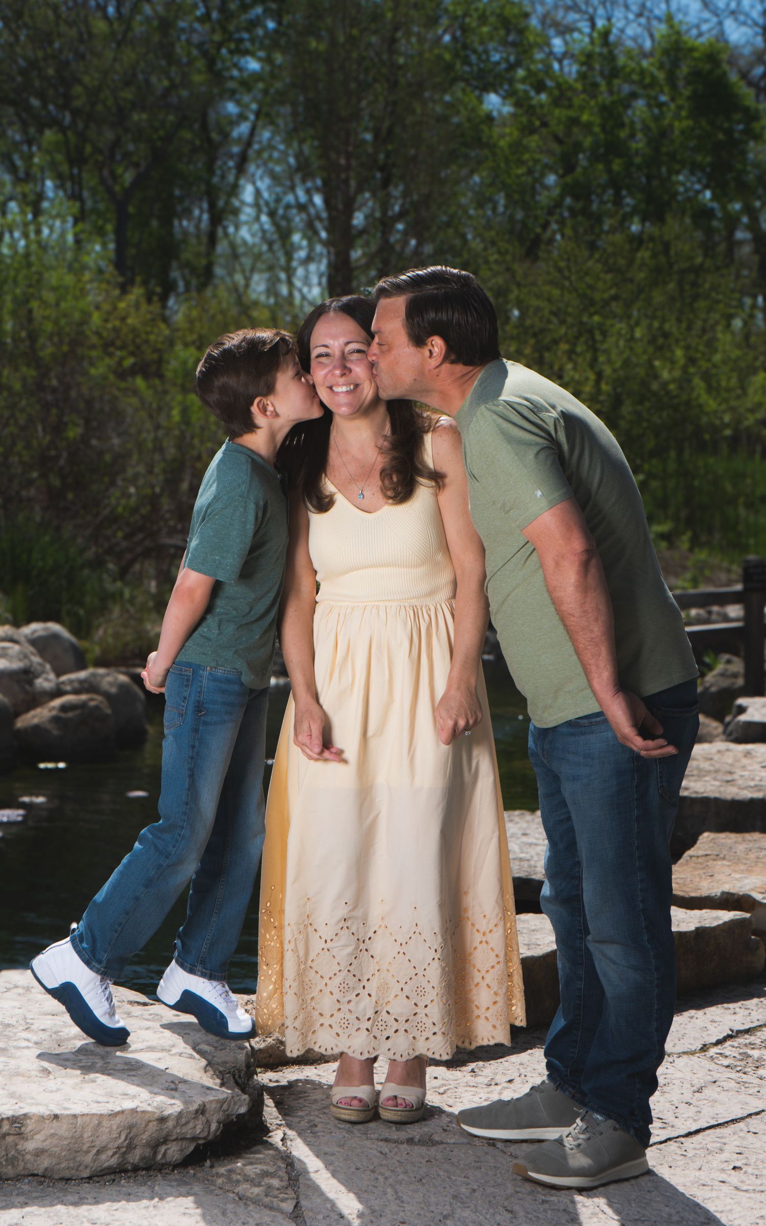 A woman in a yellow dress smiles as a boy and man kiss her cheeks outdoors near a pond.