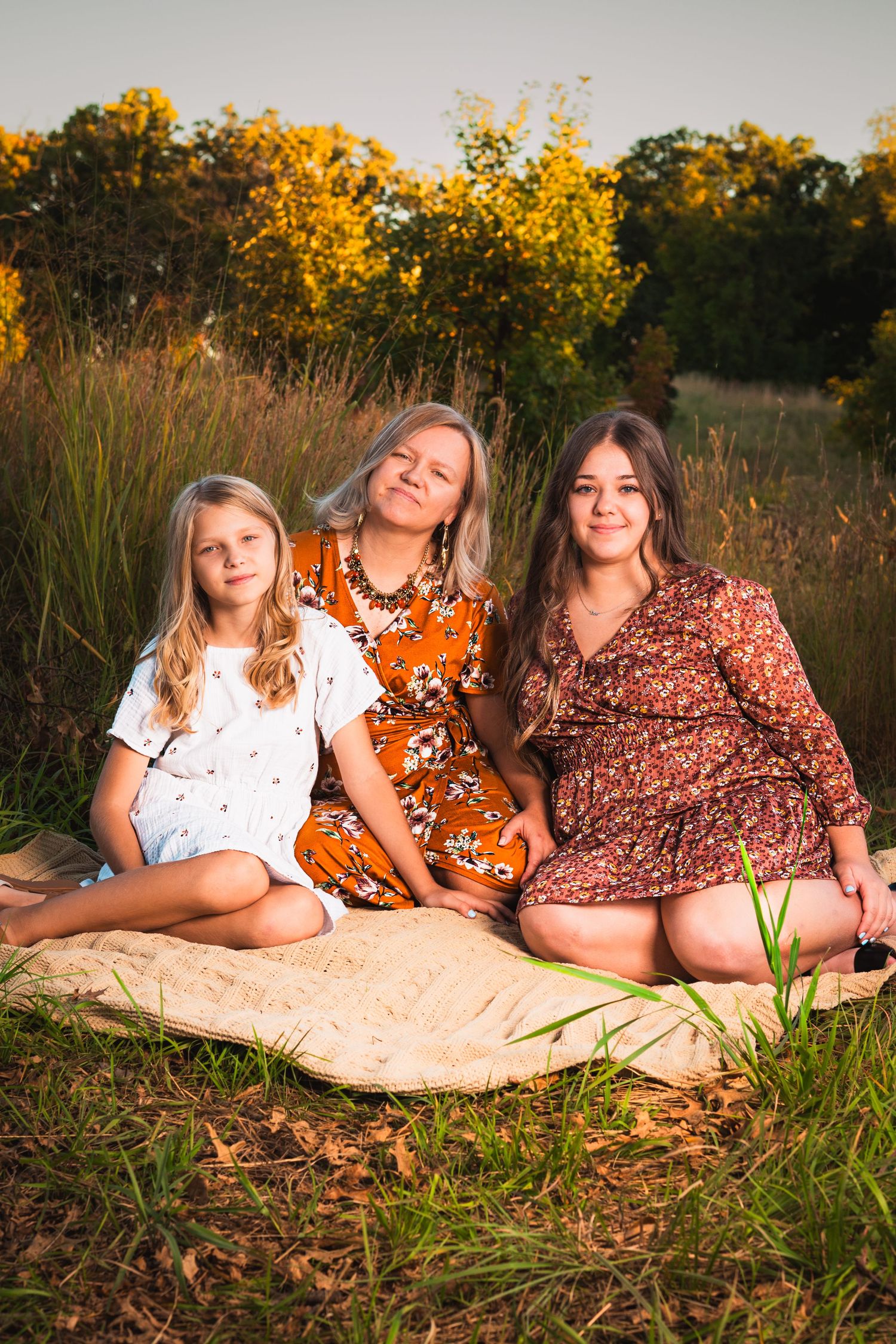 Three smiling girls in floral and white dresses sit together on a tan blanket in a grassy field at golden hour.