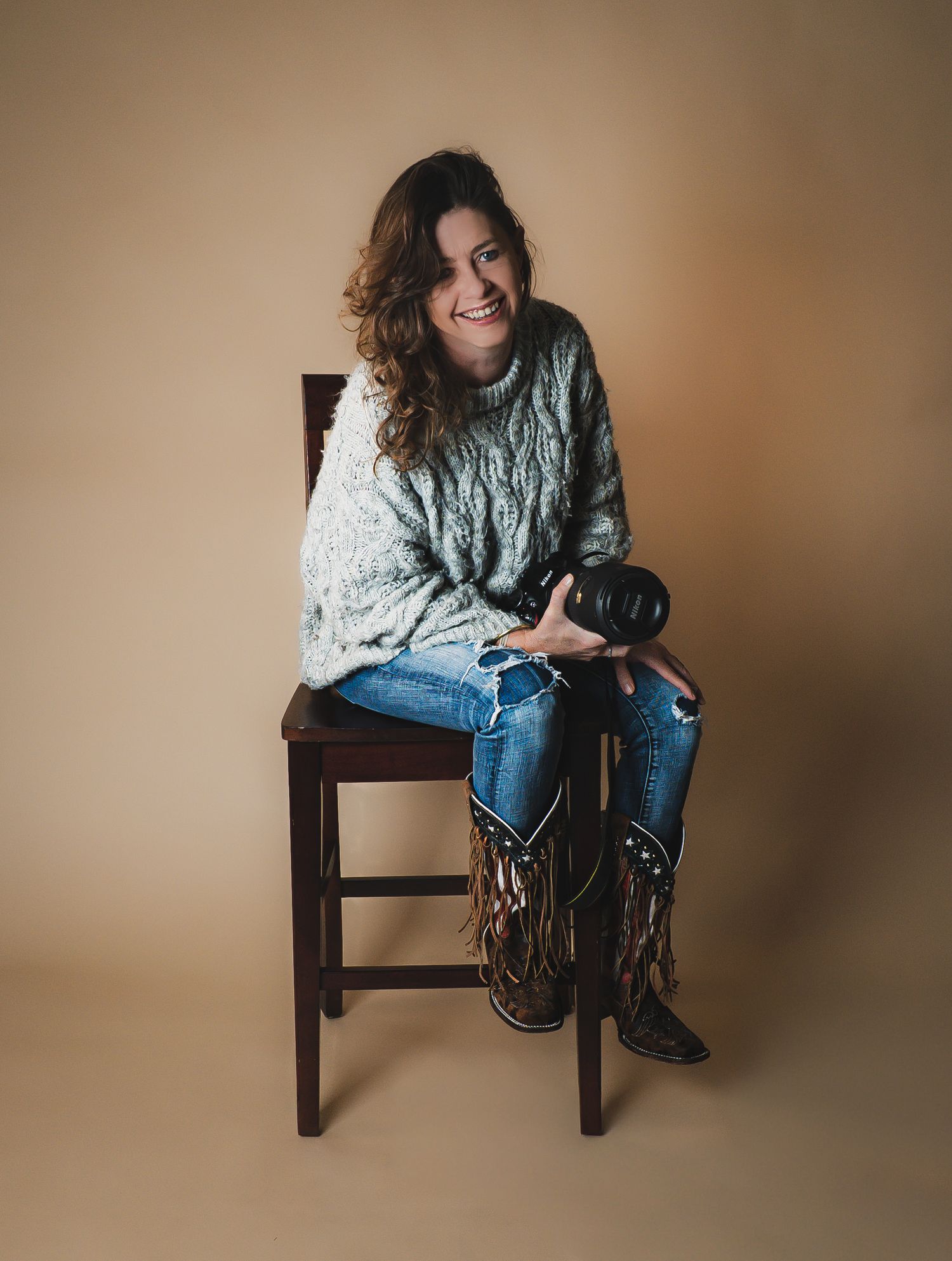 A person wearing a chunky knit sweater and jeans sits casually on a wooden chair against a neutral beige backdrop.