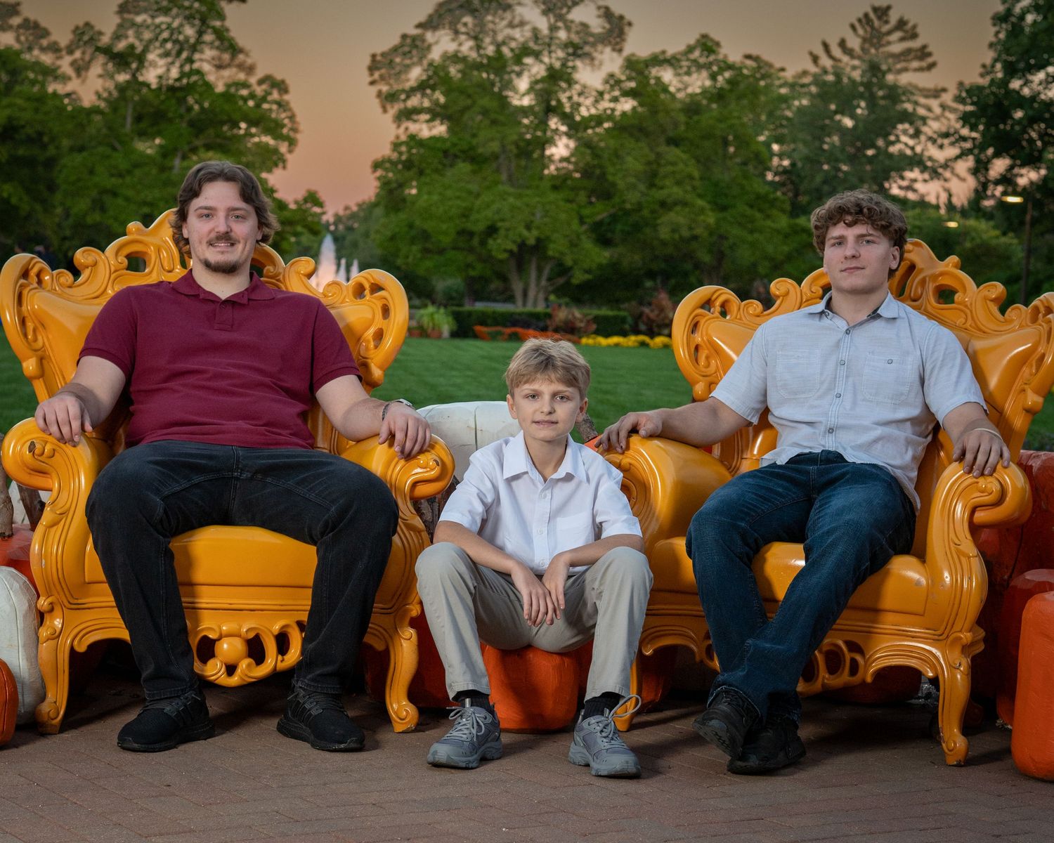 Two young men and a smiling boy sit on golden orange baroque chairs in a park at dusk, surrounded by trees and pumpkins.