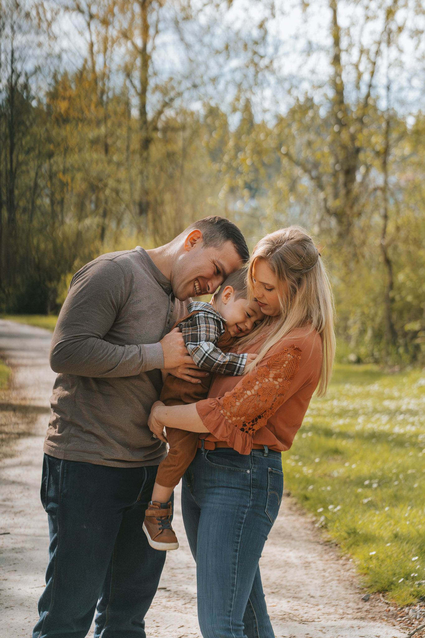 A happy young family with a toddler shares a warm embrace on a sunny outdoor path surrounded by lush green trees.