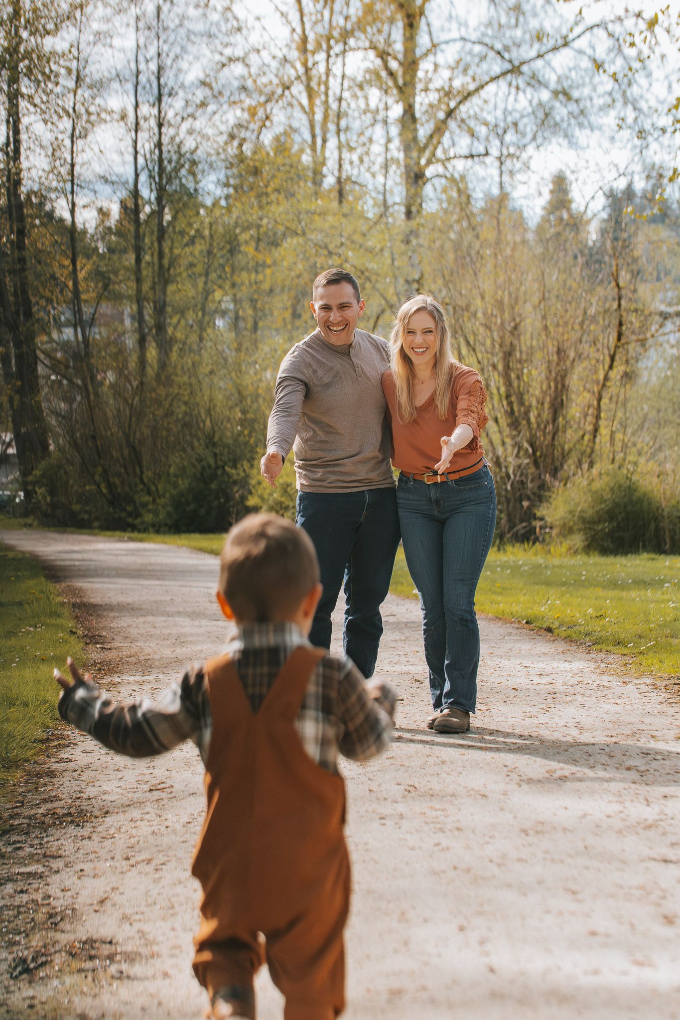 A happy couple runs toward their toddler on a sunny park path surrounded by autumn trees.
