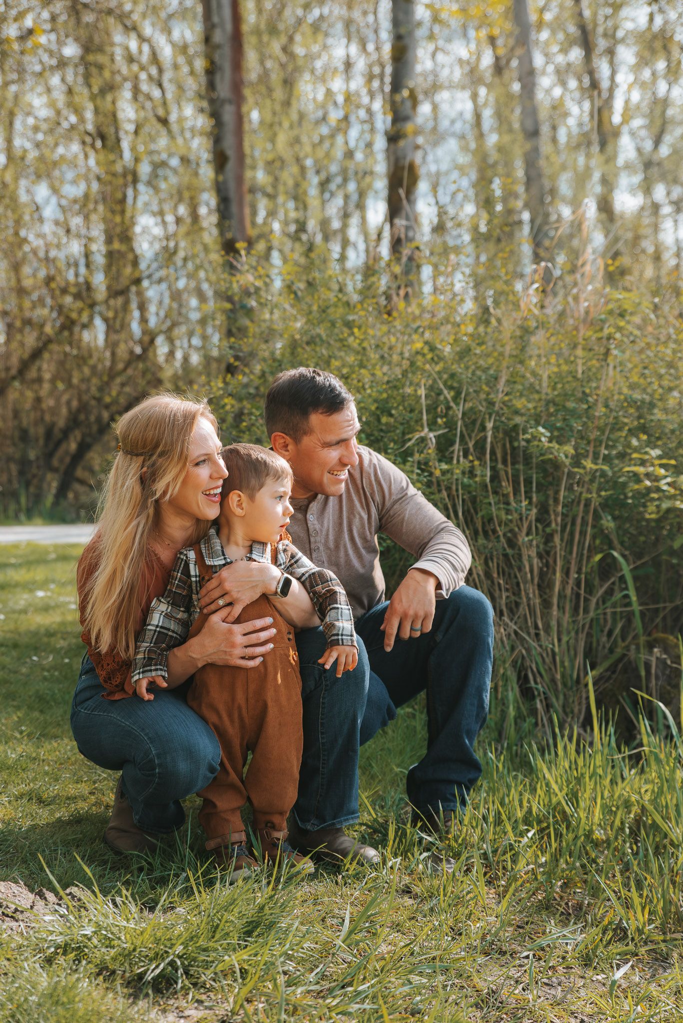 A happy family of three crouching together outdoors in a park, smiling and pointing at nature on a sunny day.