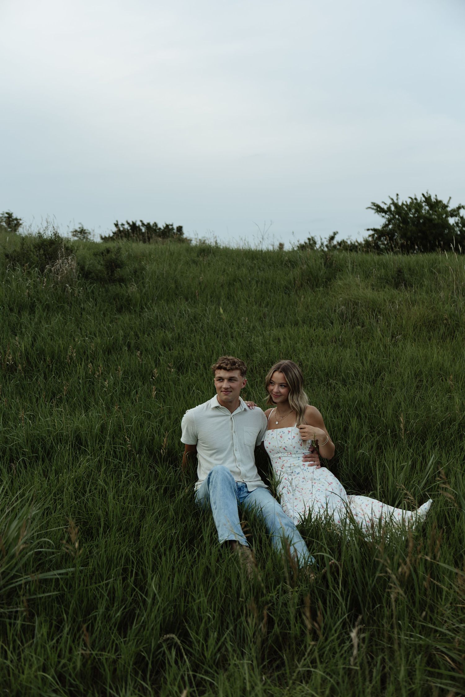 A young couple sits together in a lush green field, smiling at the camera on a cloudy day.