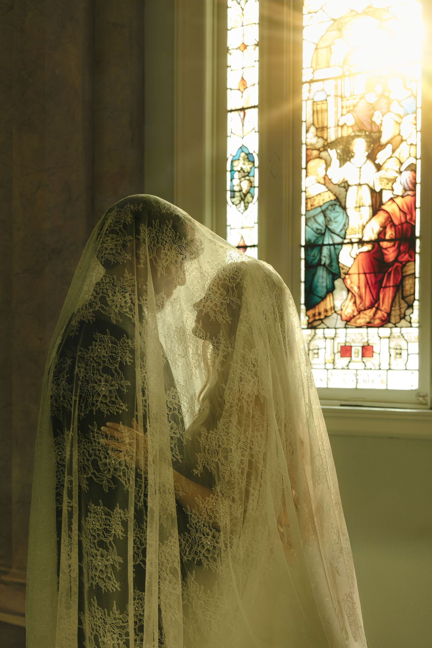 Bride and groom sharing an intimate moment under a lace veil near a stained glass church window with sunlight streaming in.