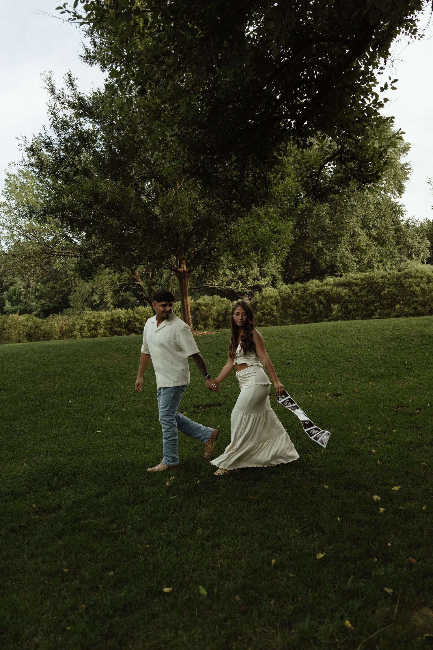 Couple in white outfits holding hands while walking on lush green grass near trees outdoors.