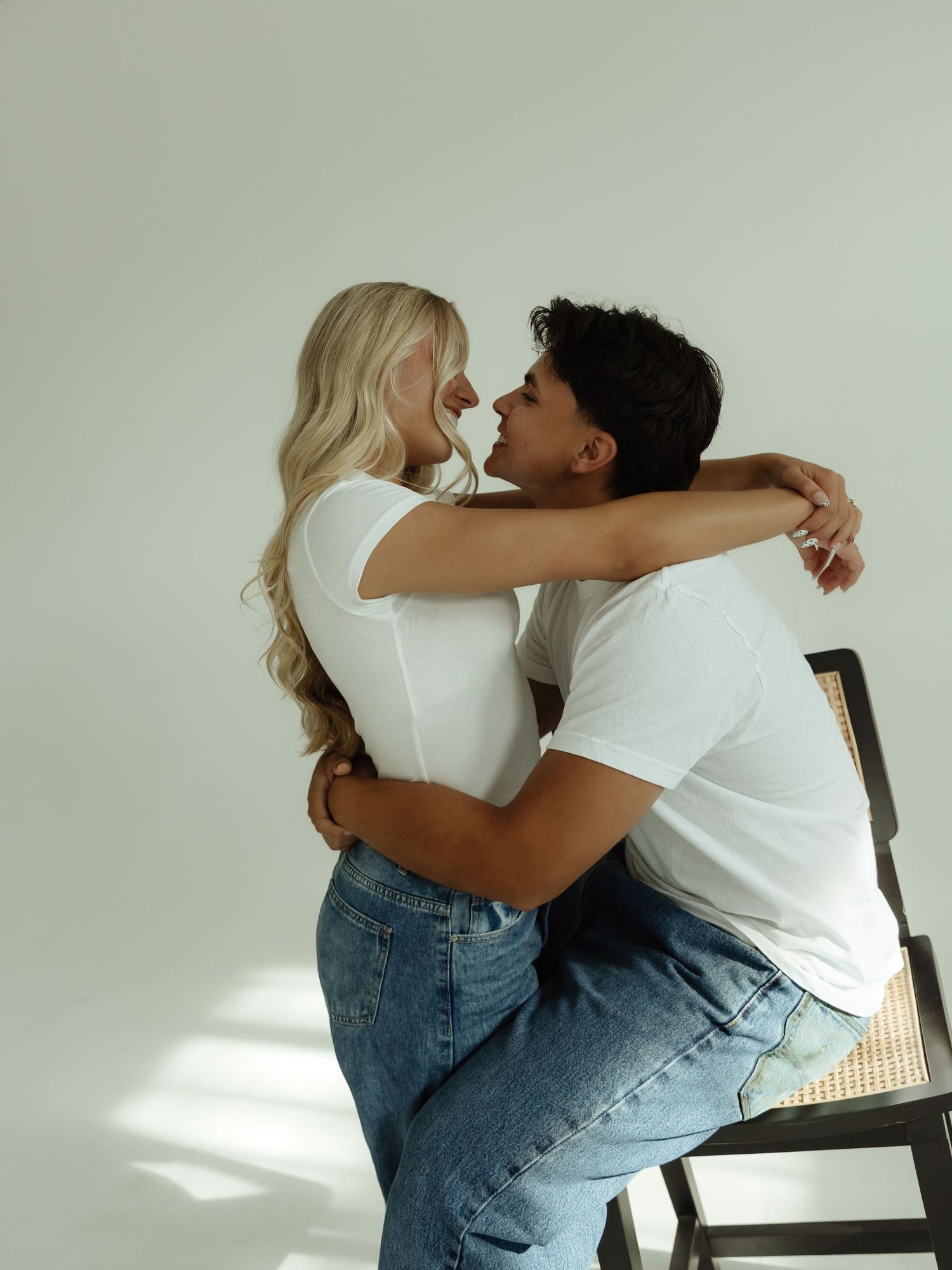 A couple in white t-shirts and jeans share a romantic embrace, with the woman sitting on a man's lap on a chair.
