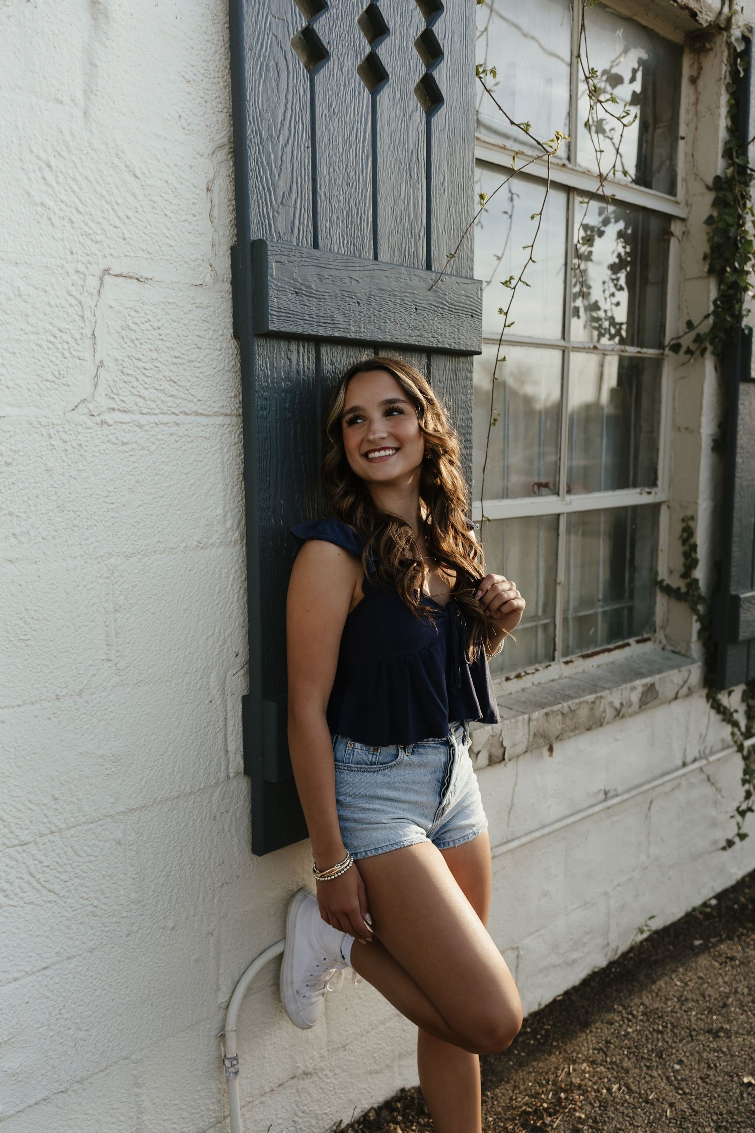 Smiling young woman in denim shorts and navy top leaning against white brick wall near vintage window.