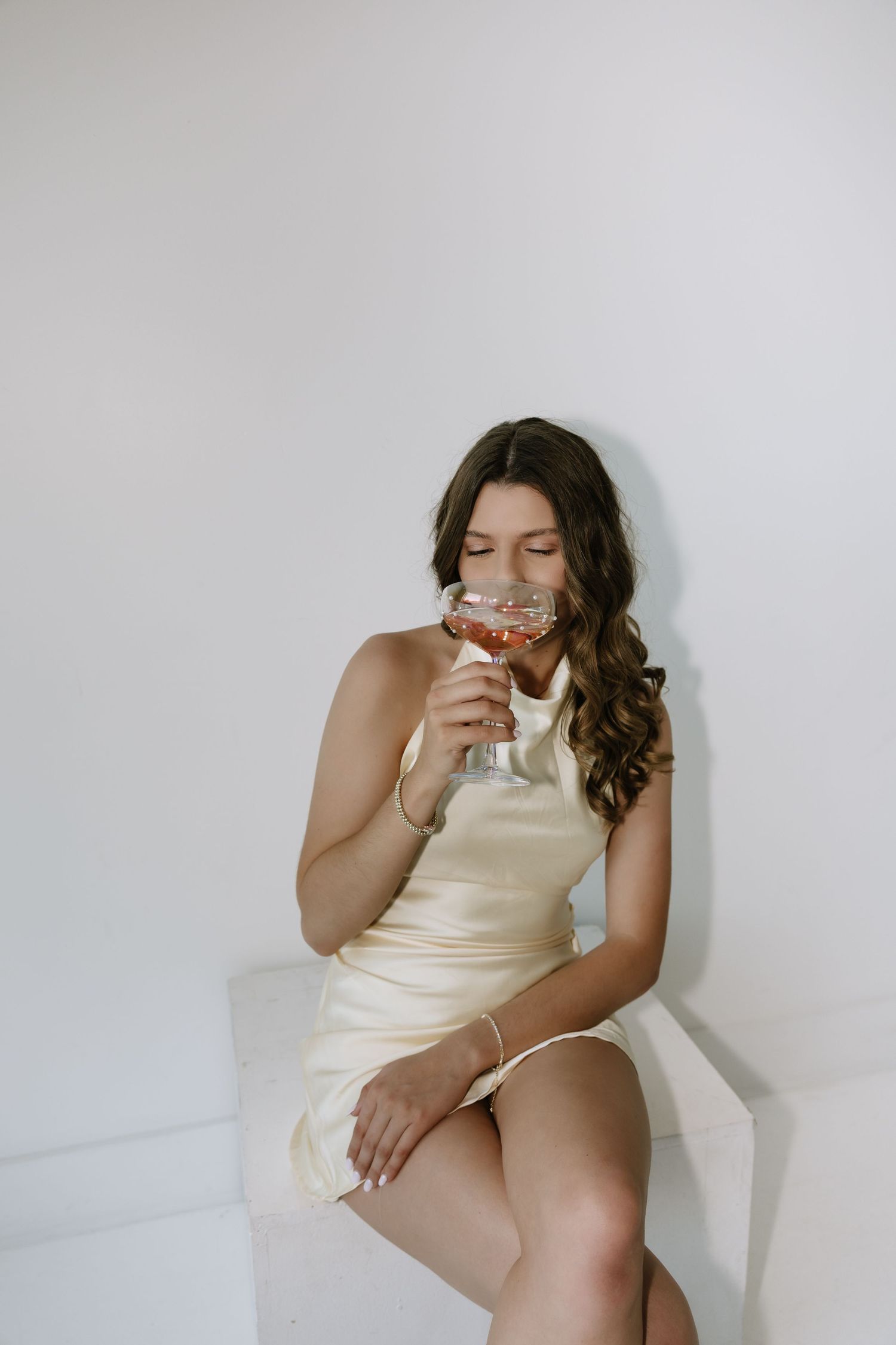Young woman in cream dress sipping rose wine from elegant glass while seated against white wall.