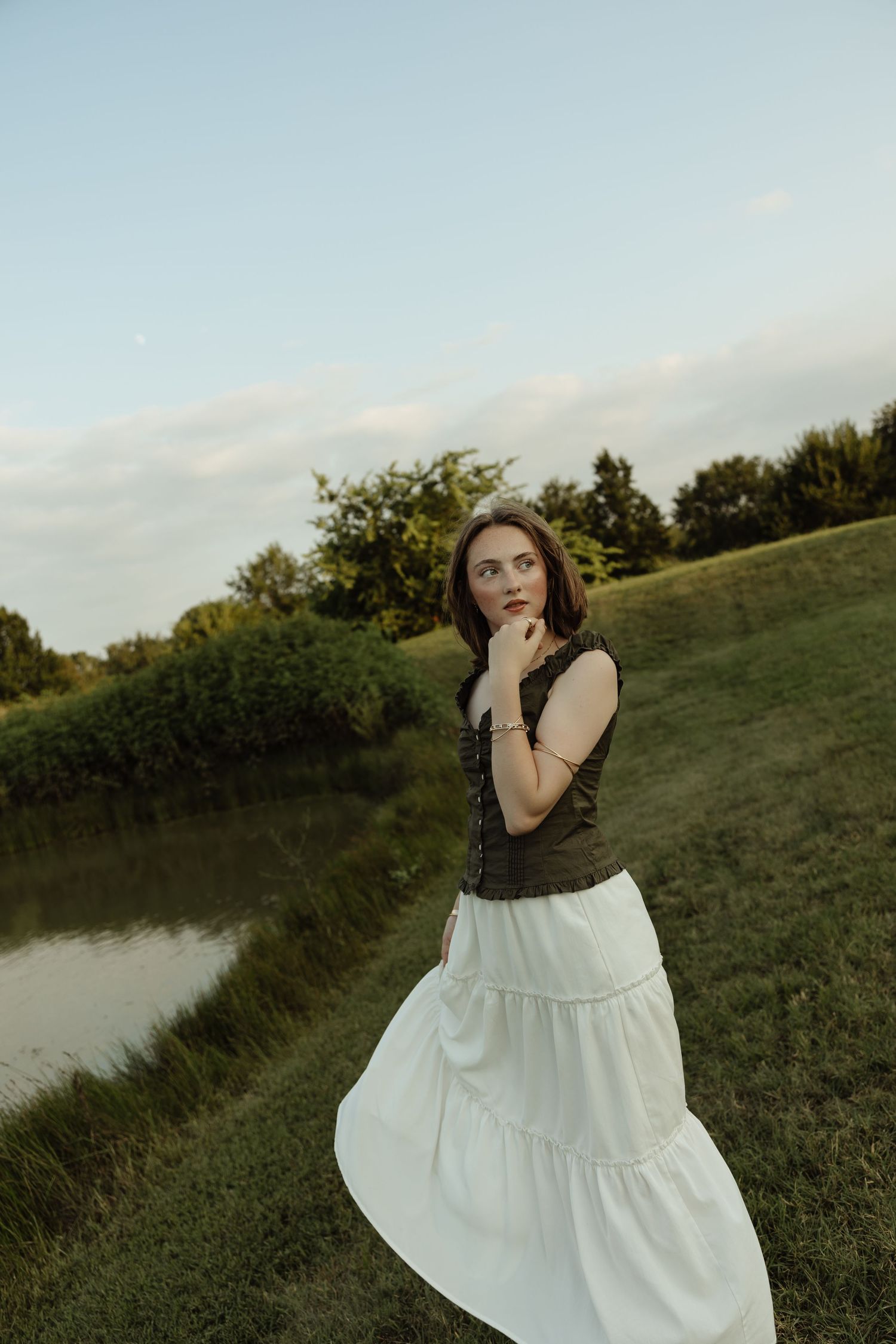 Young woman in white tiered skirt and dark top poses thoughtfully near a pond in a lush green landscape.