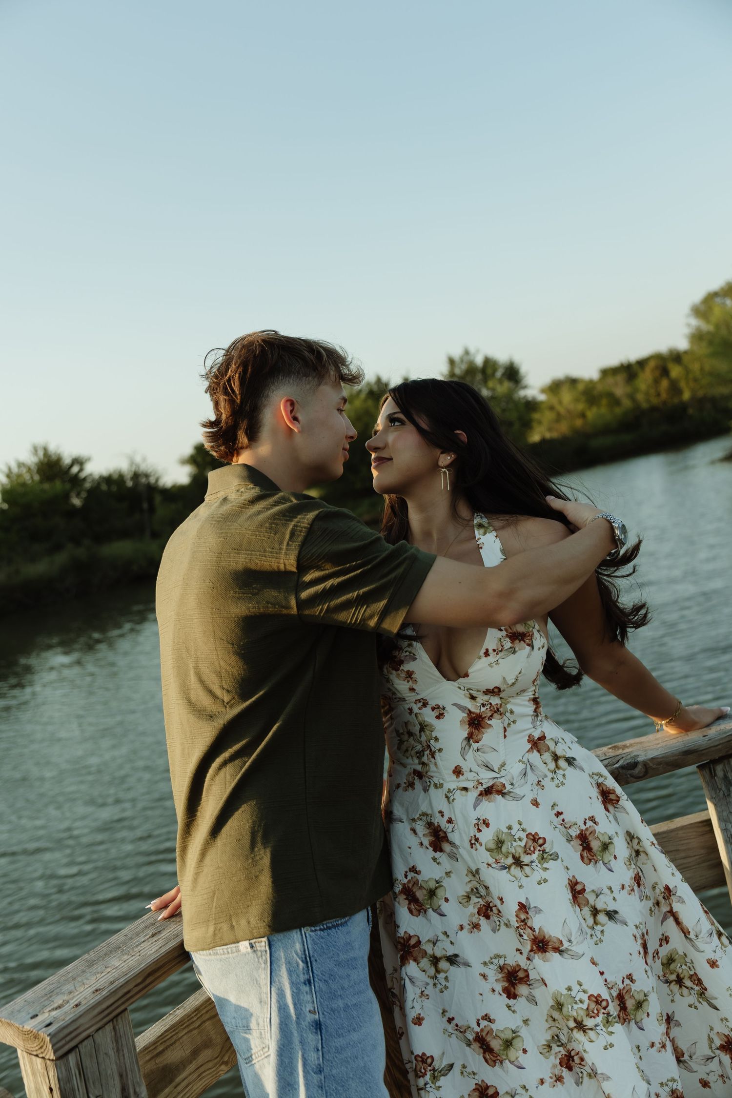 Couple sharing a romantic moment on a wooden dock by a lake, woman in floral dress, man in olive shirt.