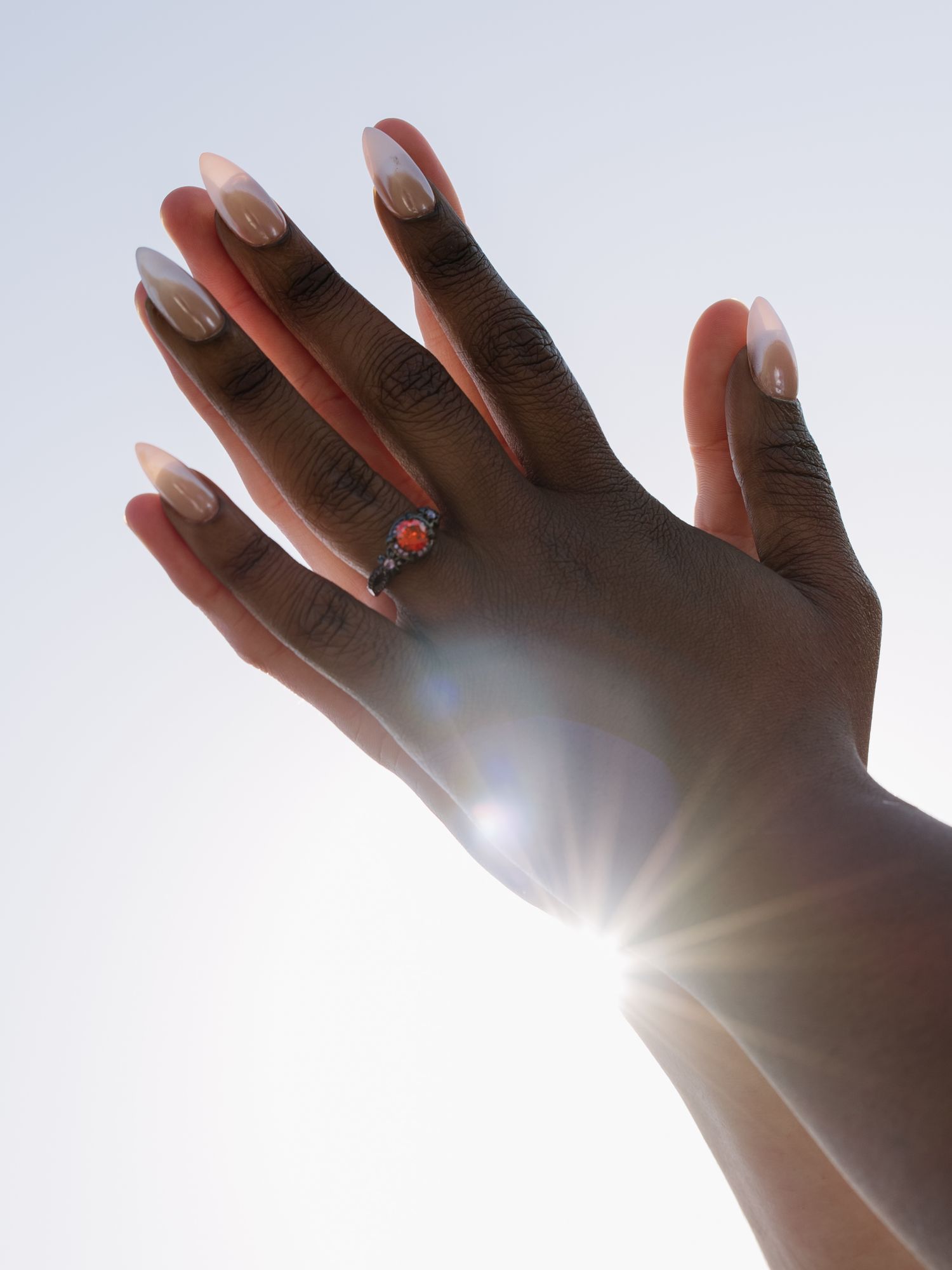 Hand with French tip nails and orange gemstone ring raised against bright sunlit sky.