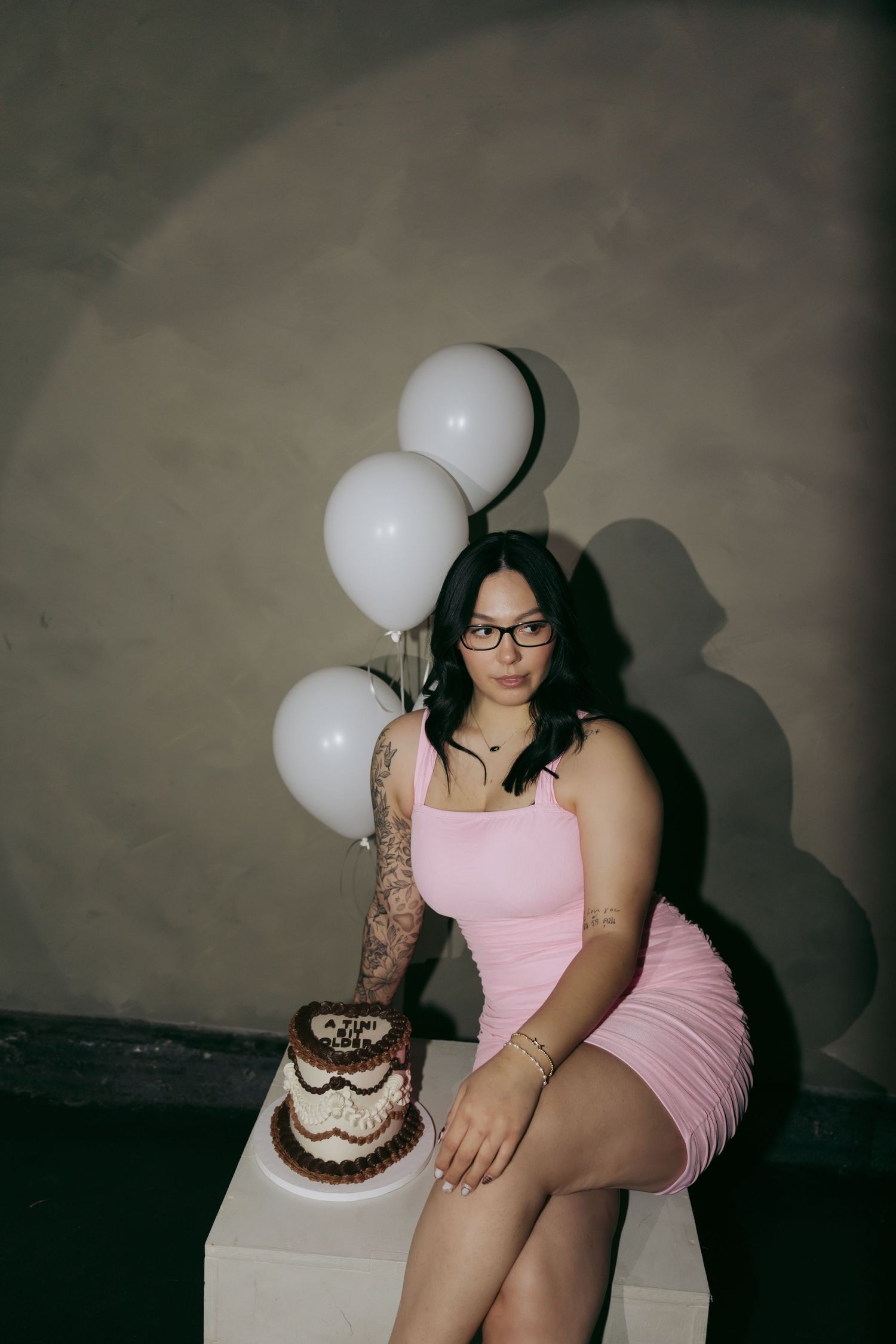 Woman in pink dress with tattoos poses beside birthday cake and white balloons against gray wall.