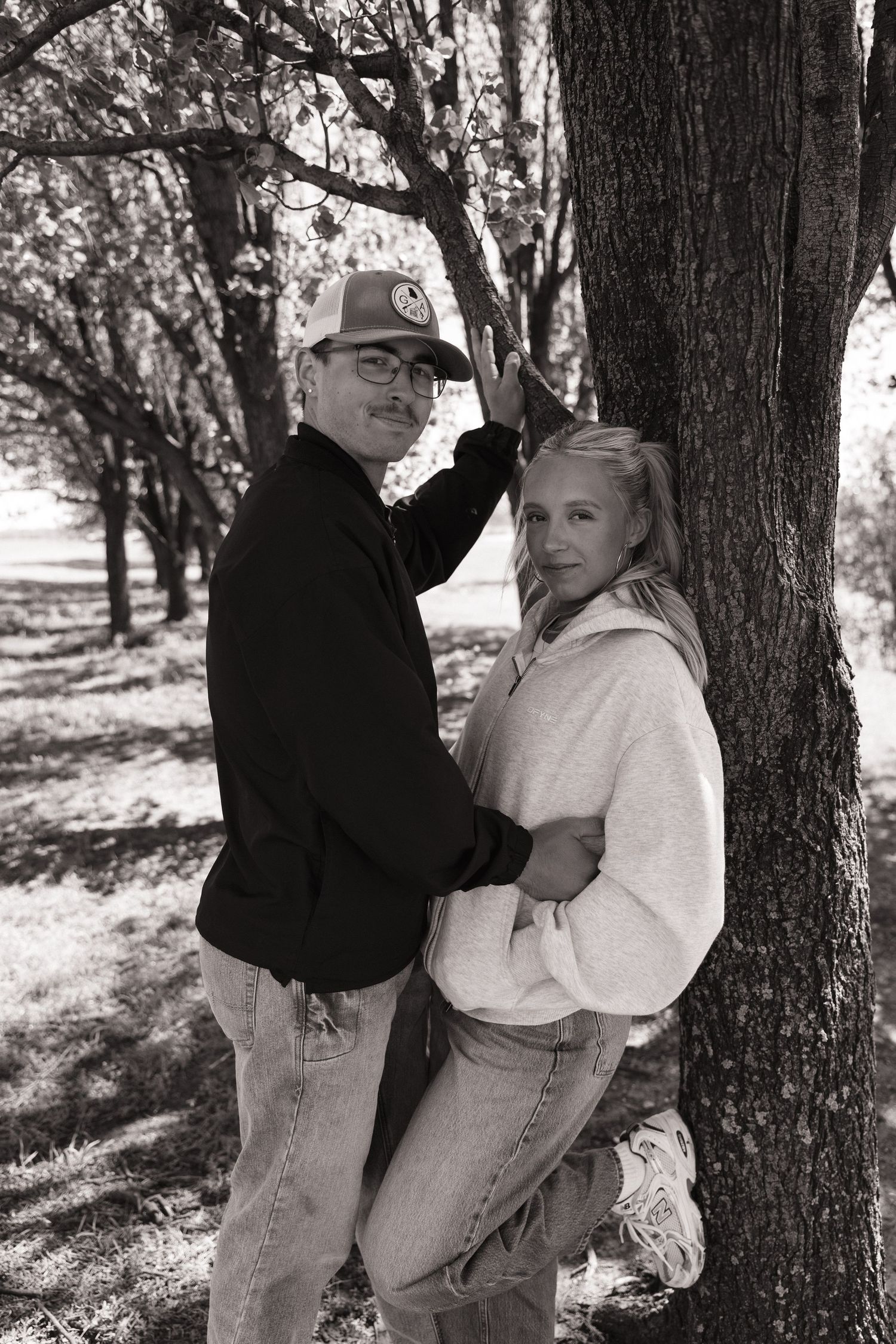 Black and white photo of a couple posing by a tree outdoors, man in cap and jacket, woman in hoodie and jeans.
