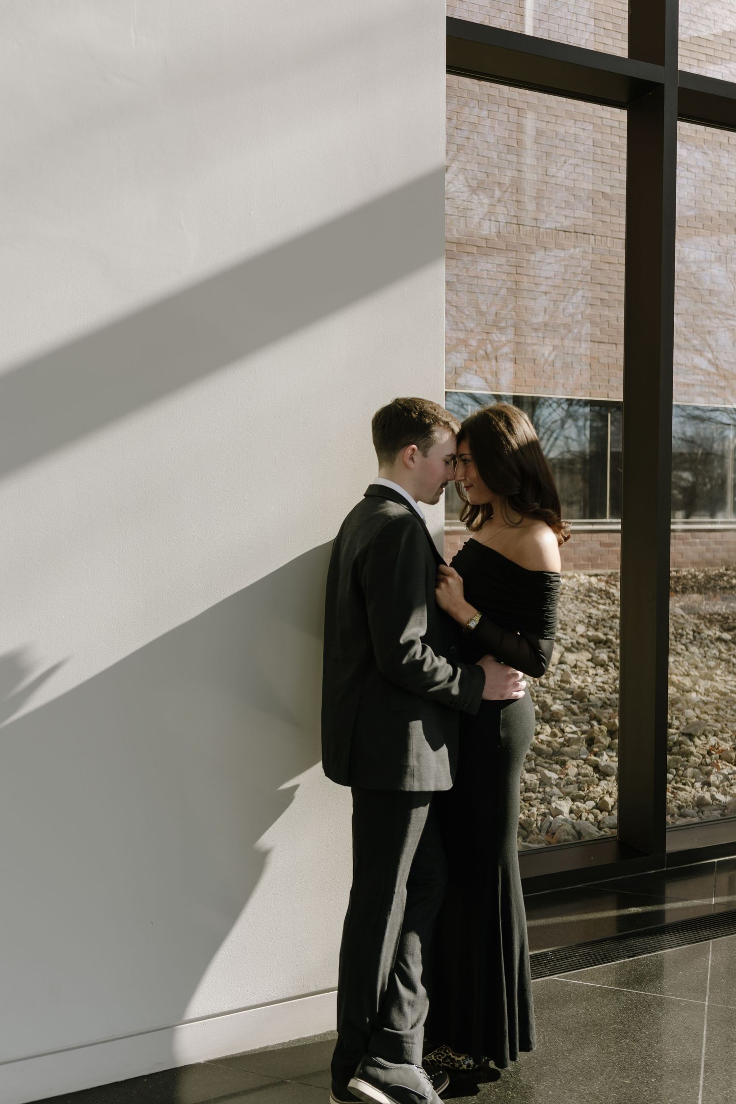 Couple in elegant black attire sharing a romantic moment by a sunlit window in a modern building.