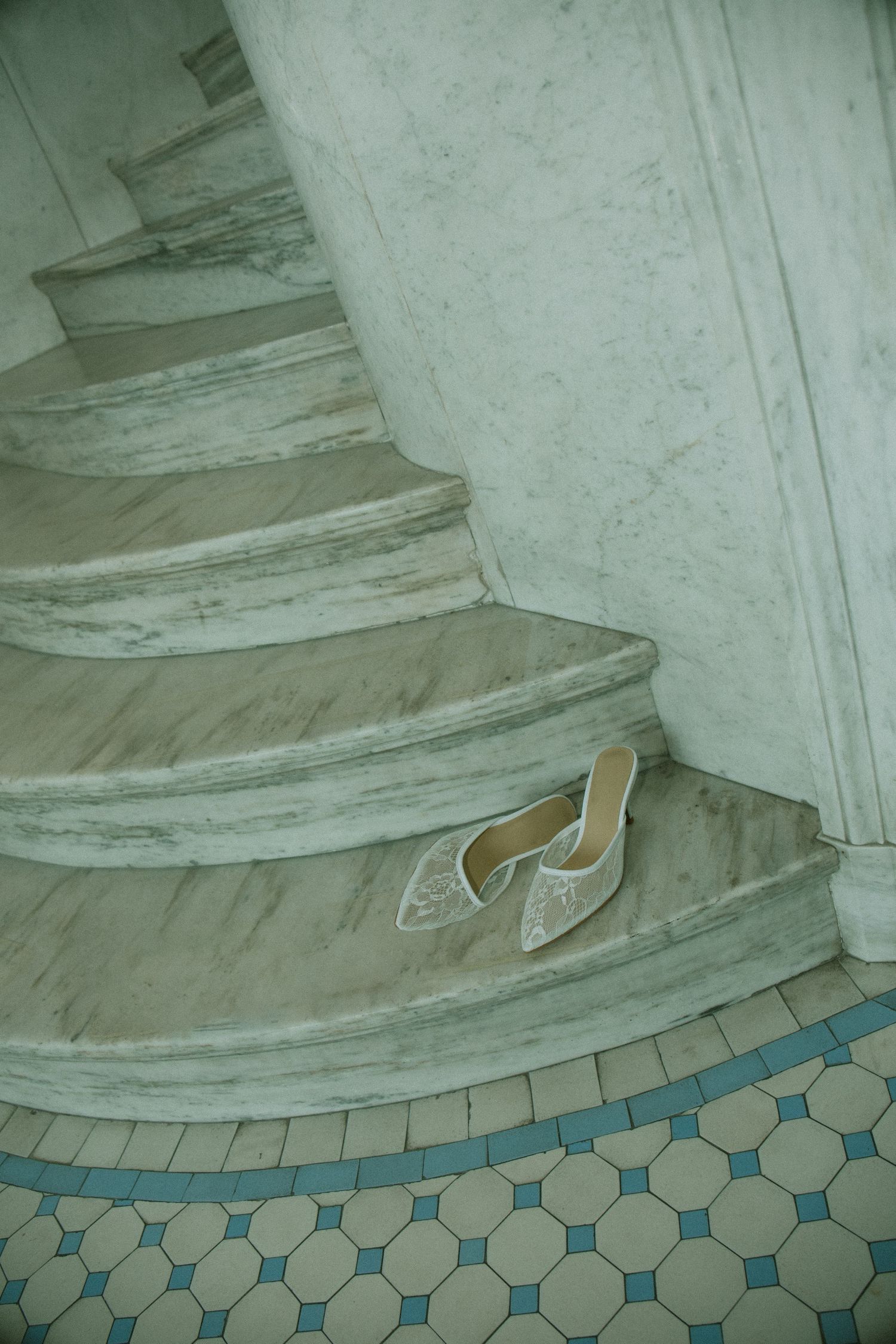 Elegant white lace bridal heels resting on curved marble stairs with ornate tiled floor.