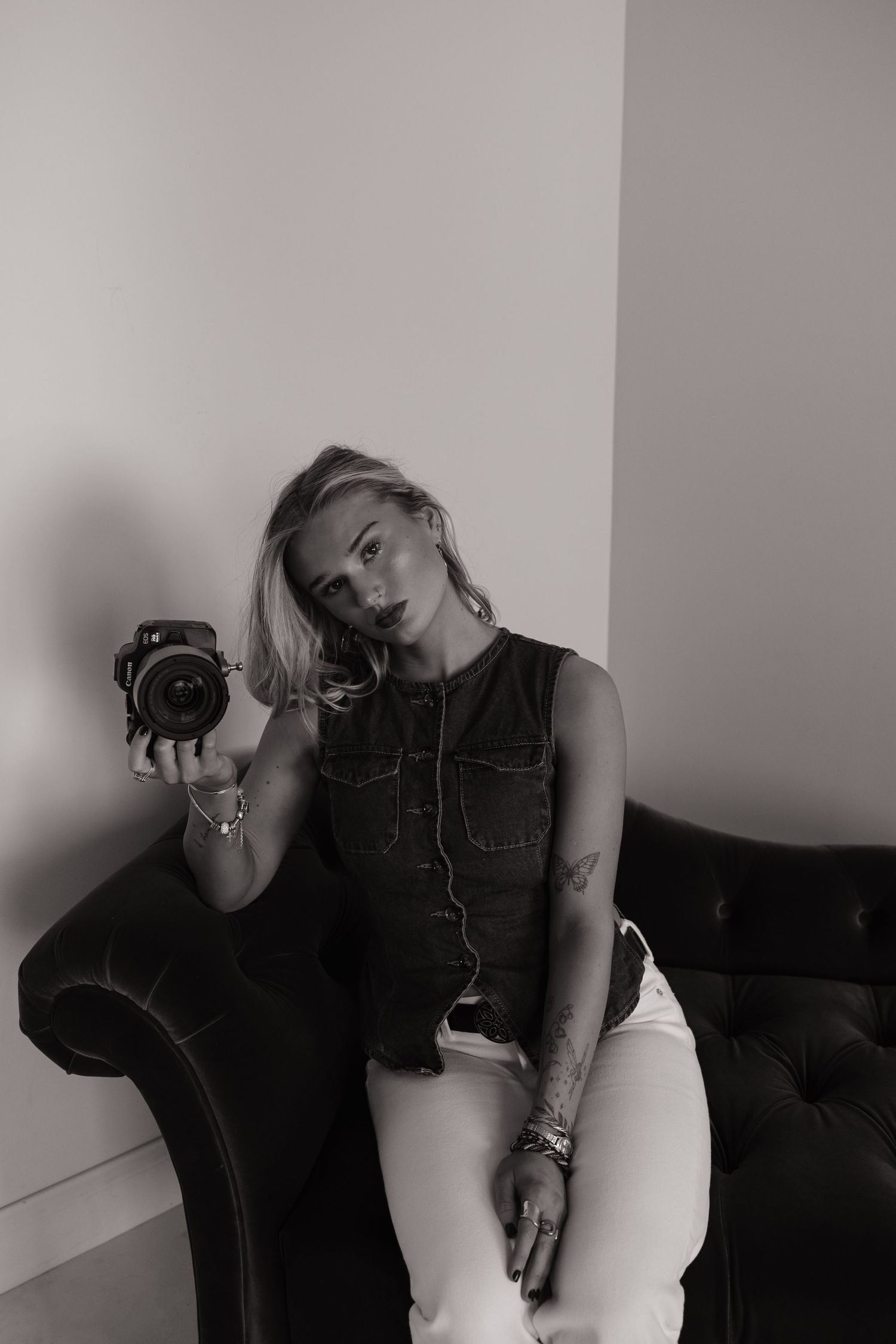 Black and white photo of a blonde woman sitting on a tufted sofa holding a camera, wearing a denim vest and white jeans.