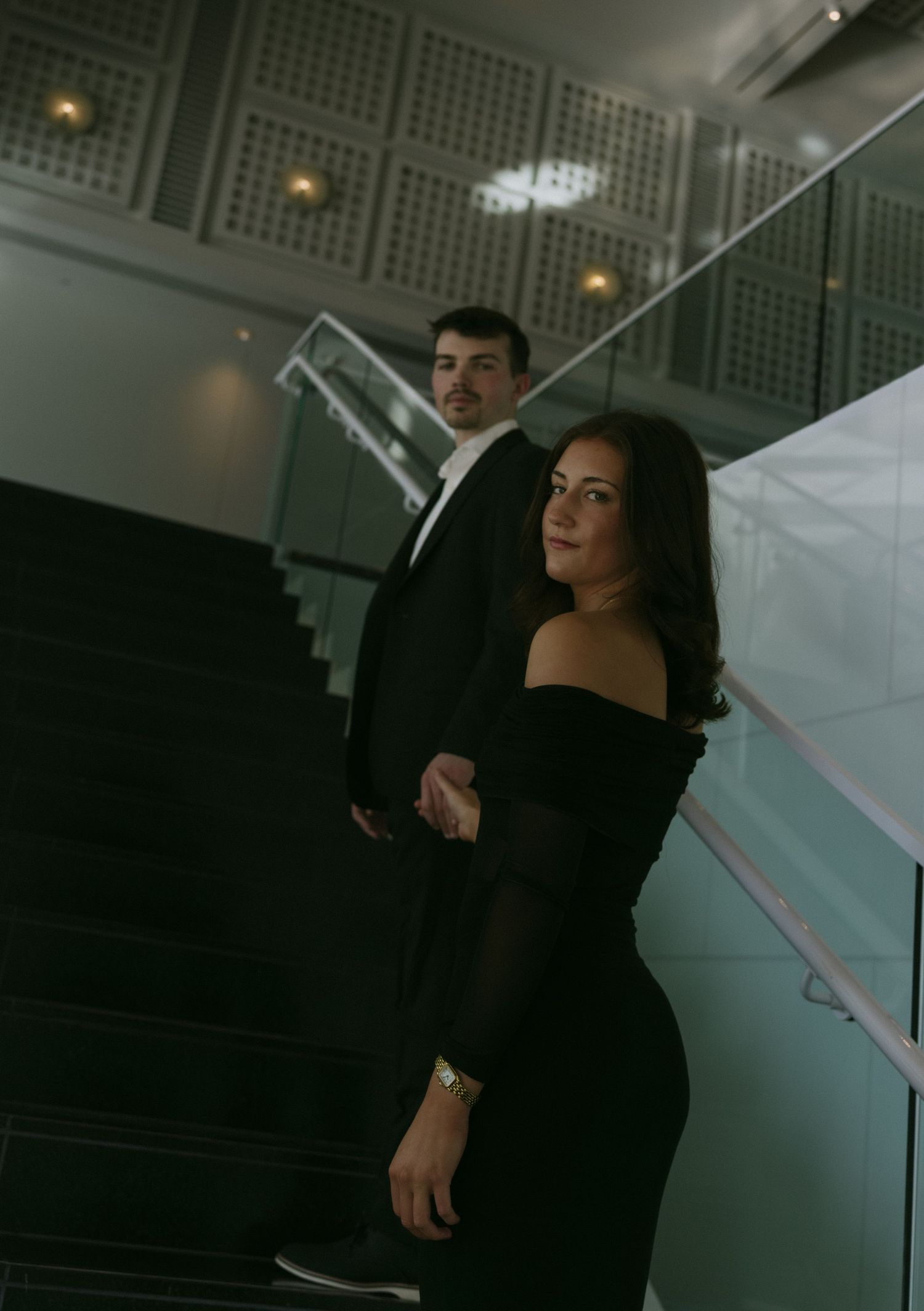 Elegant couple in black formal attire posing on a staircase, woman in off-shoulder dress, man in tuxedo behind her.