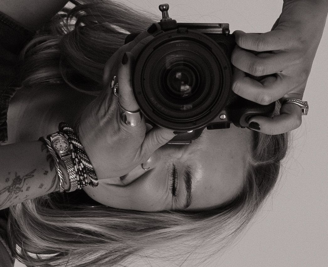 Black and white photo of a woman holding a camera up to her face, wearing bracelets and rings.