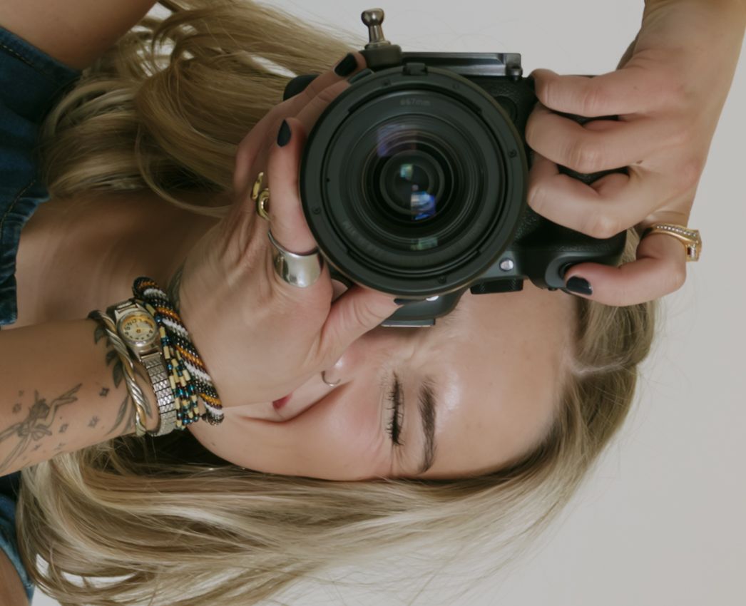 A blonde woman holds a large camera up to her face, wearing gold bracelets and a tattoo on her wrist.