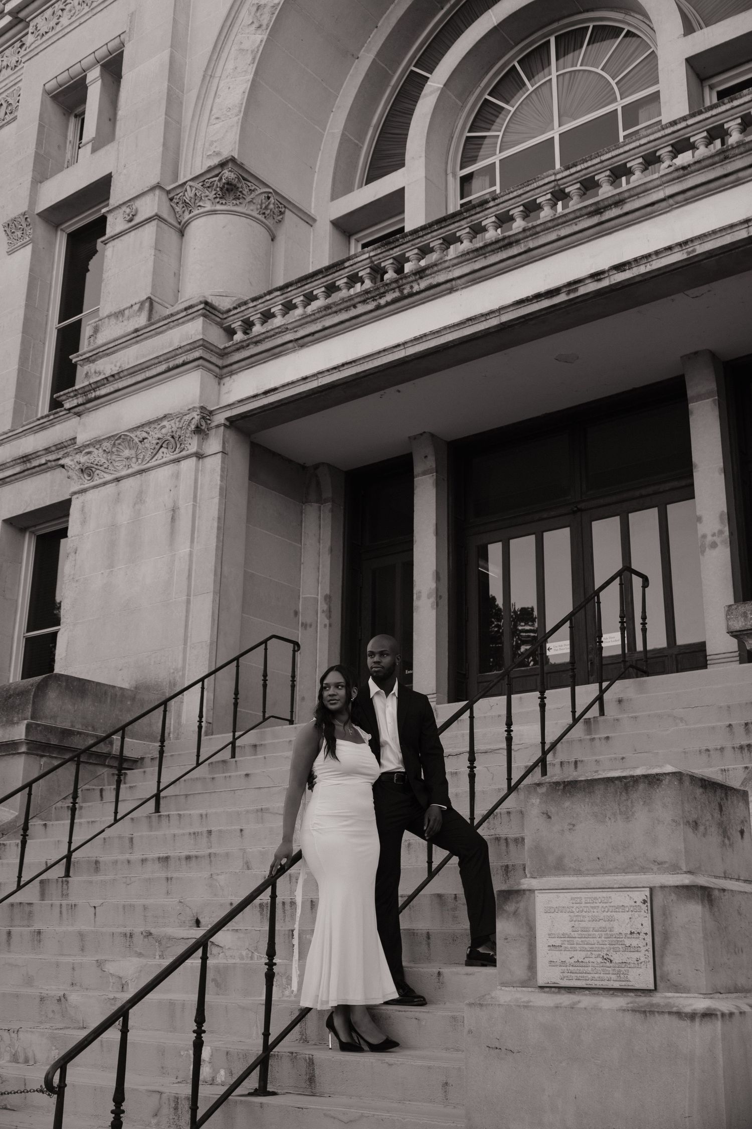Black and white photo of a couple in formal attire posing on steps of a grand classical building.